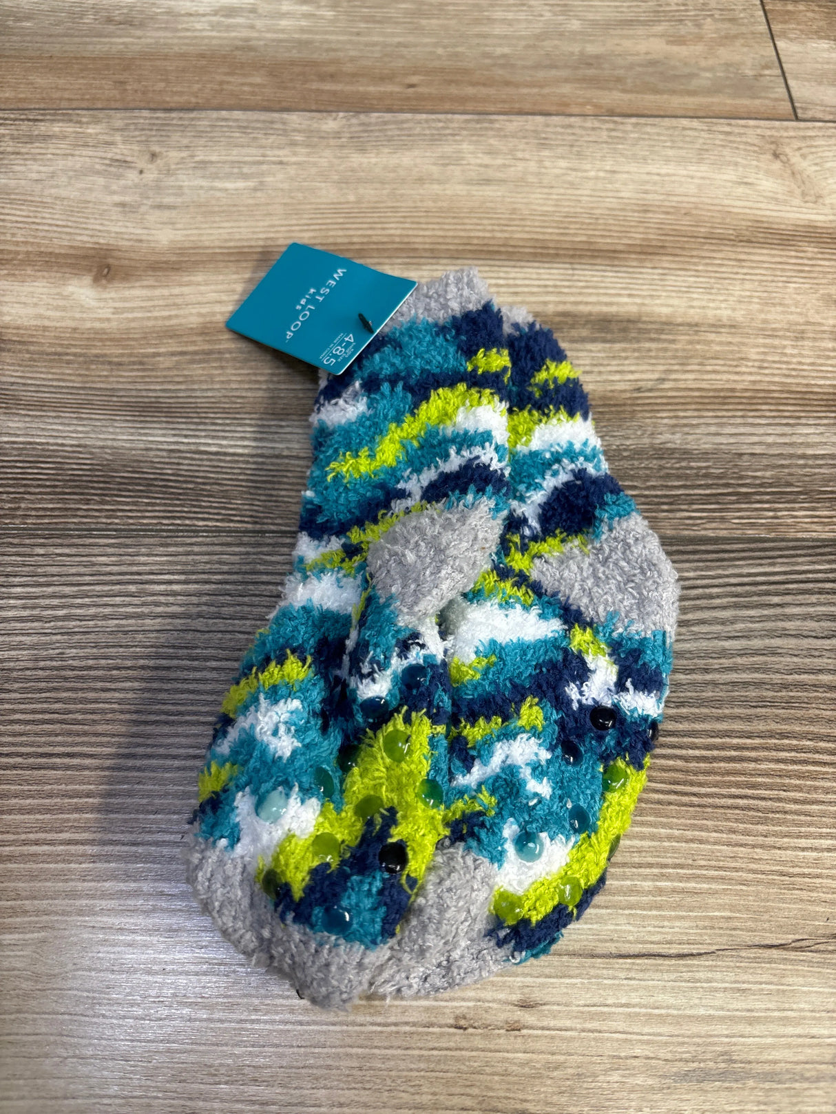 Top-down view of a kids fuzzy sock showing non-slip gripper dots and blue teal lime gray stripes on a wood floor