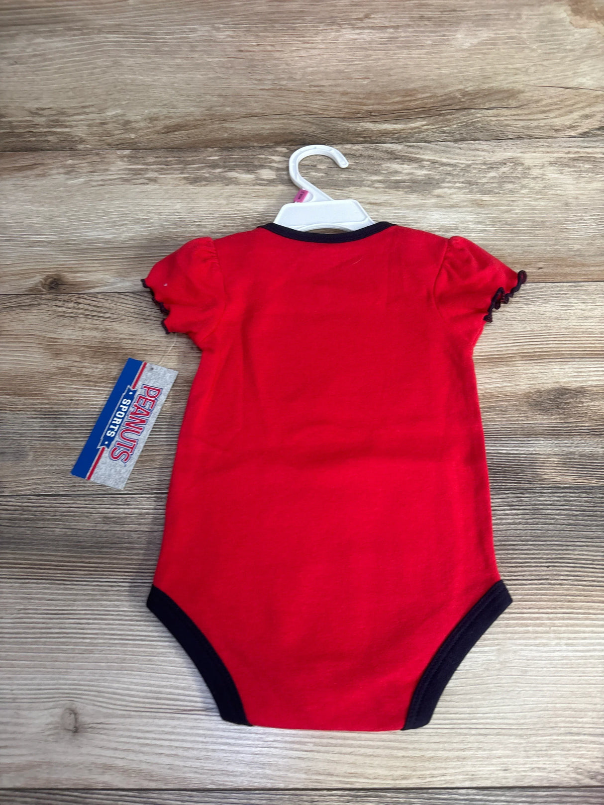 Red baby bodysuit with black trim on hanger, Peanuts Sports tag, wood background
