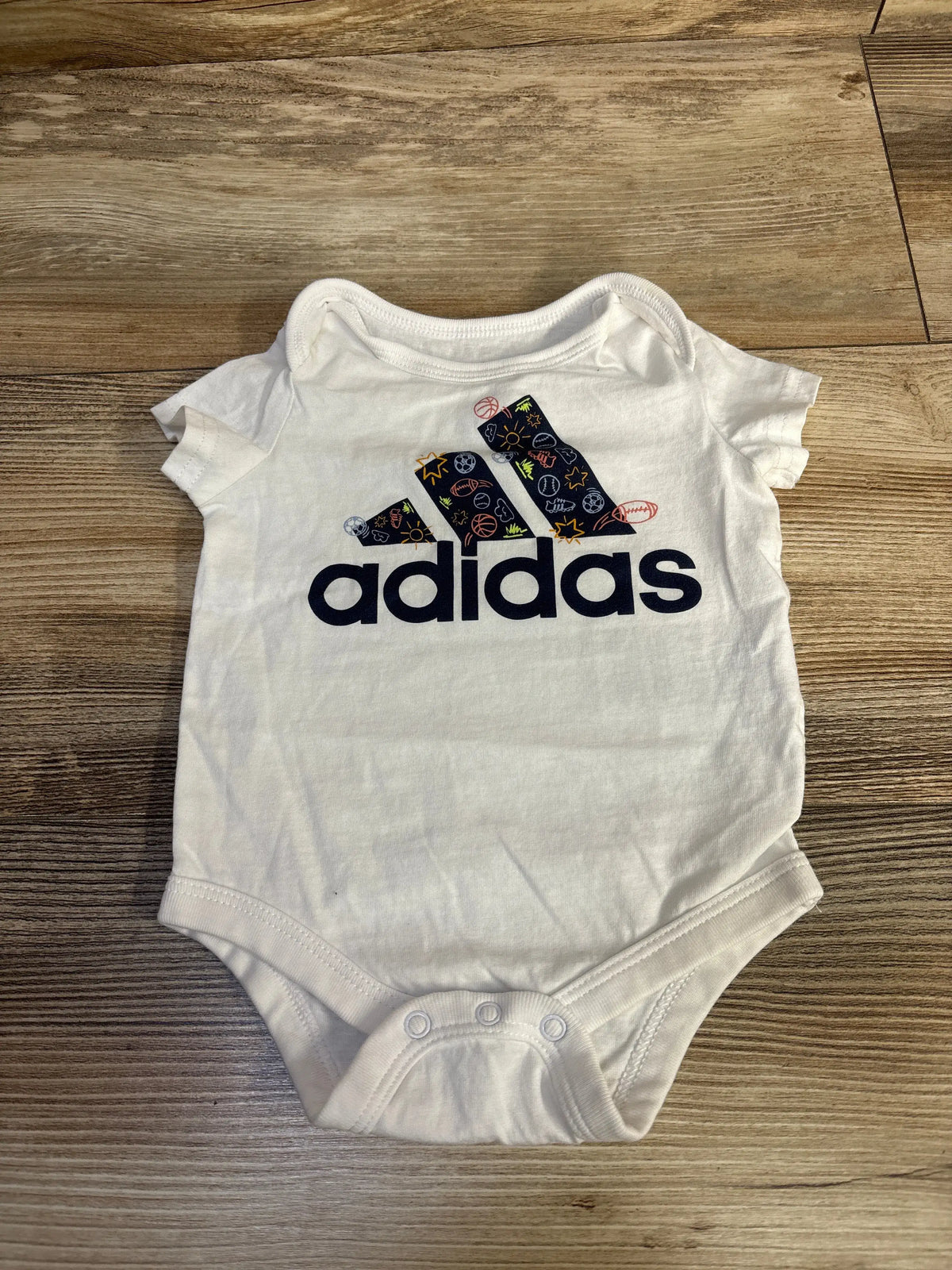 Front view of Adidas white baby bodysuit, size 3 months, showcasing a colorful sports-ball Adidas logo on a wood floor