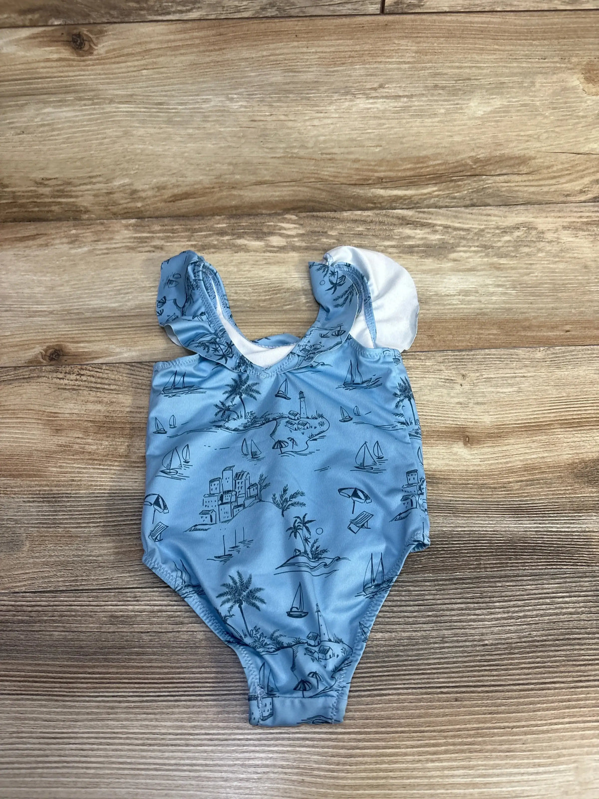 Flat lay of blue Zara baby one-piece swimsuit showing beach print and ruffle shoulder straps on wood background, size 6 to 12 months