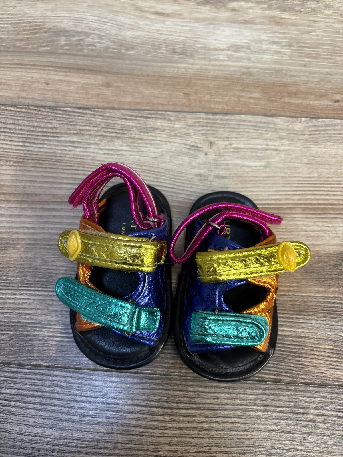 Top view of Kurt Geiger London Orson metallic toddler sandals with rainbow straps and two Velcro closures on wood floor