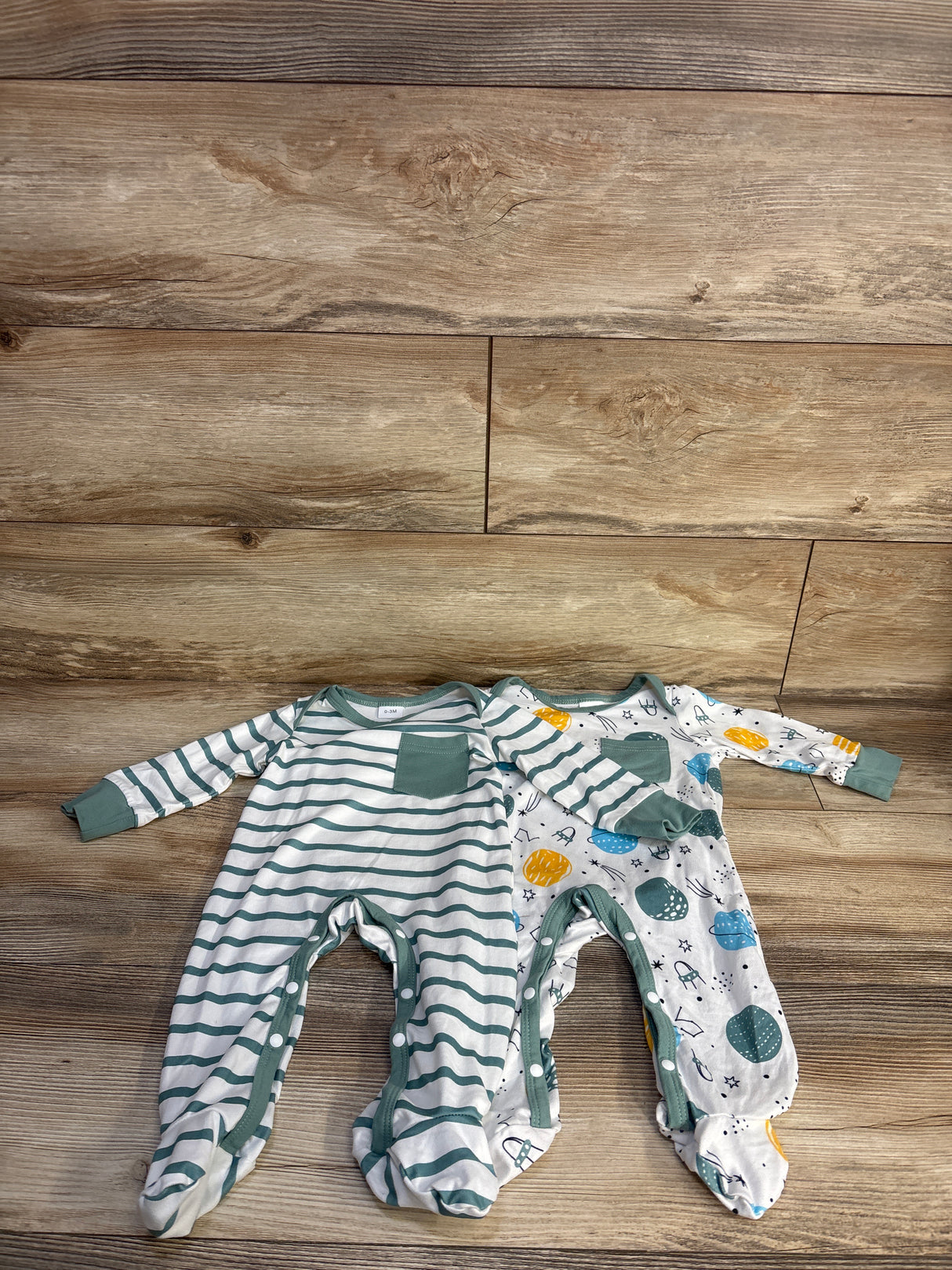 Top-down view of two newborn footed coveralls (2-pack) showcasing a green-striped coverall with chest pocket and a space-print coverall on wood background