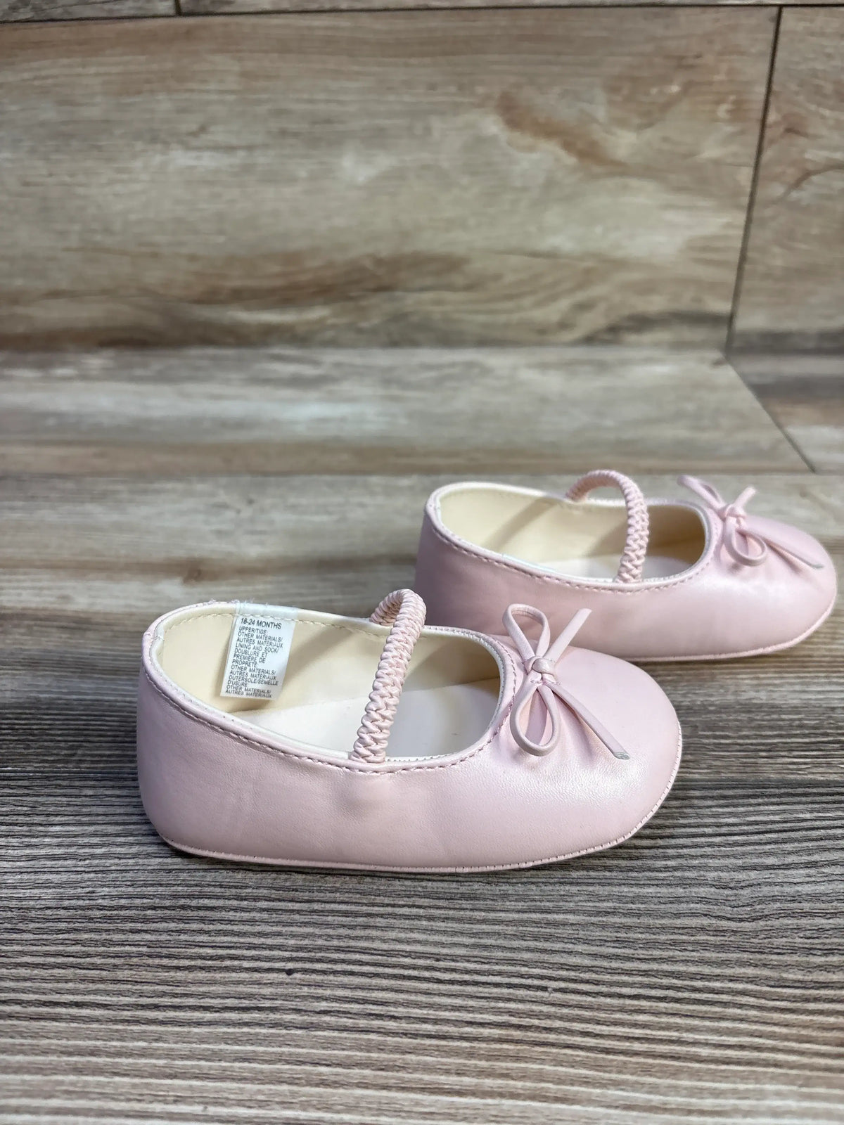 Three-quarter side view of Janie & Jack soft sole pink ballet flats showing braided elastic strap and bow on wood background