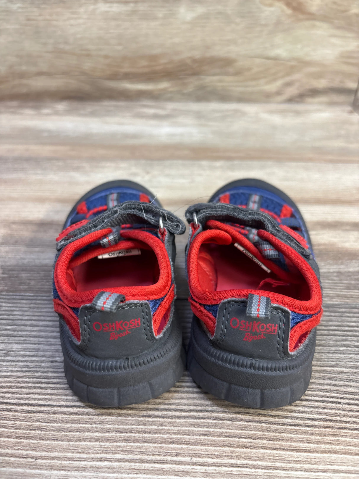 Rear view of OshKosh toddler bumptoe sandals in navy and red showing heel logo and Velcro straps on wood surface