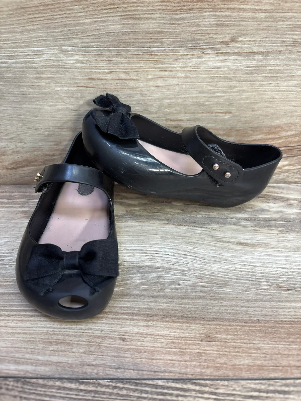 Front view of Mini Melissa Ultragirl Bow black jelly flats showing bow, Mary Jane strap, and peep-toe on light wood background