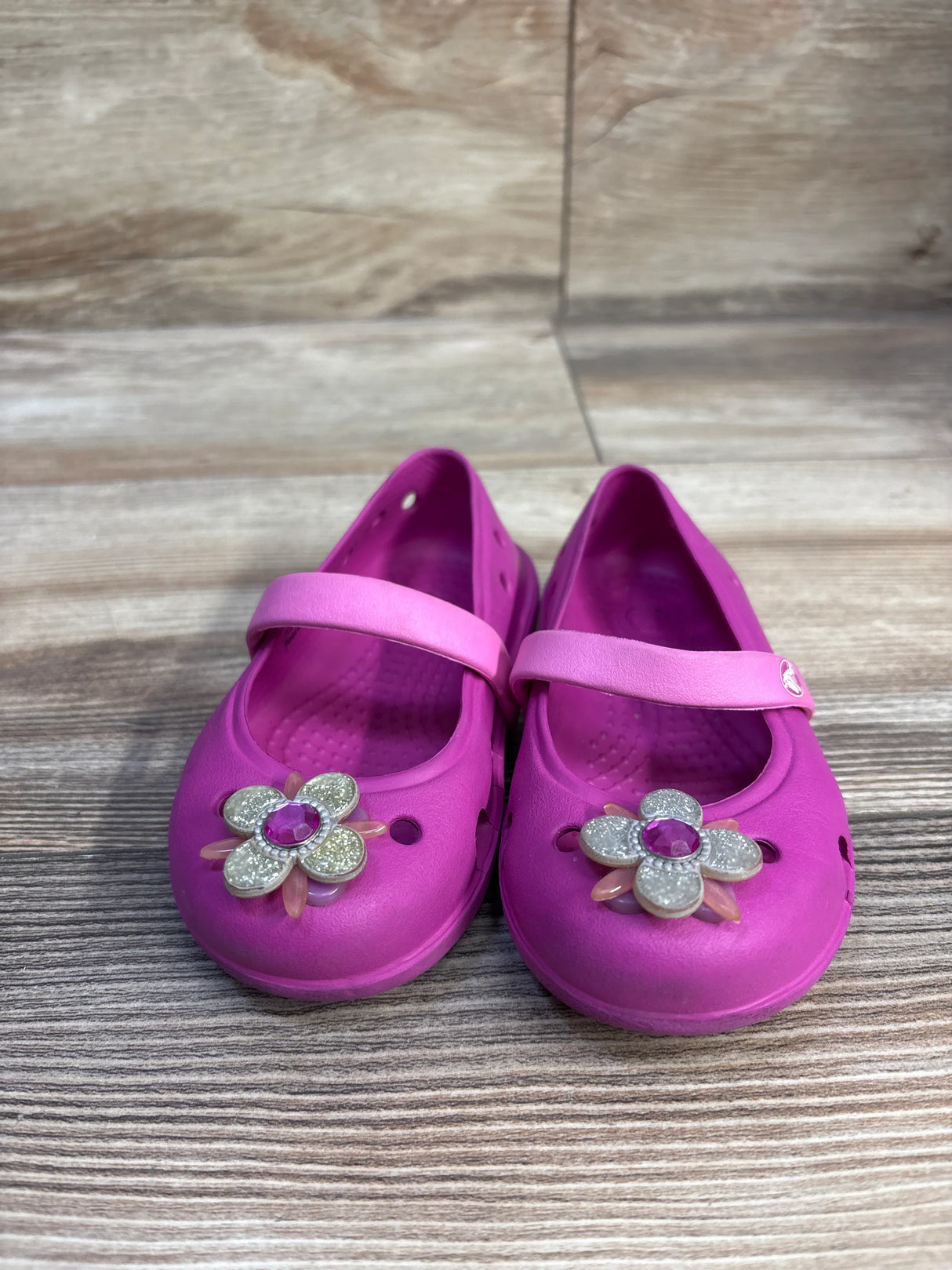 Front view of Crocs Keeley Petal Charm Flats showcasing glittery flower charms on the toes and single Mary Jane straps on a wood-grain floor