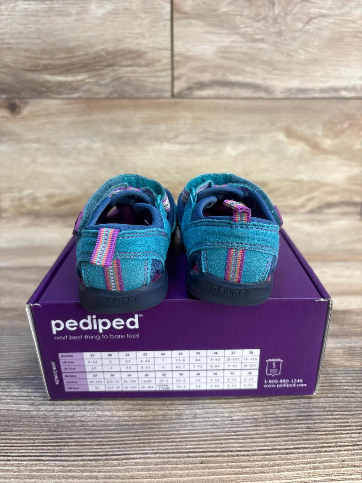 Rear view of Pediped Flex Sahara toddler sandals in blue showing striped heel tabs and Velcro straps on a purple Pediped box, wood background