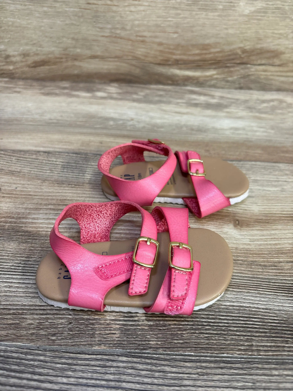 Three-quarter view of Baby Gap pink toddler buckle sandals showcasing double front straps with gold buckles and tan cushioned insoles on wood background