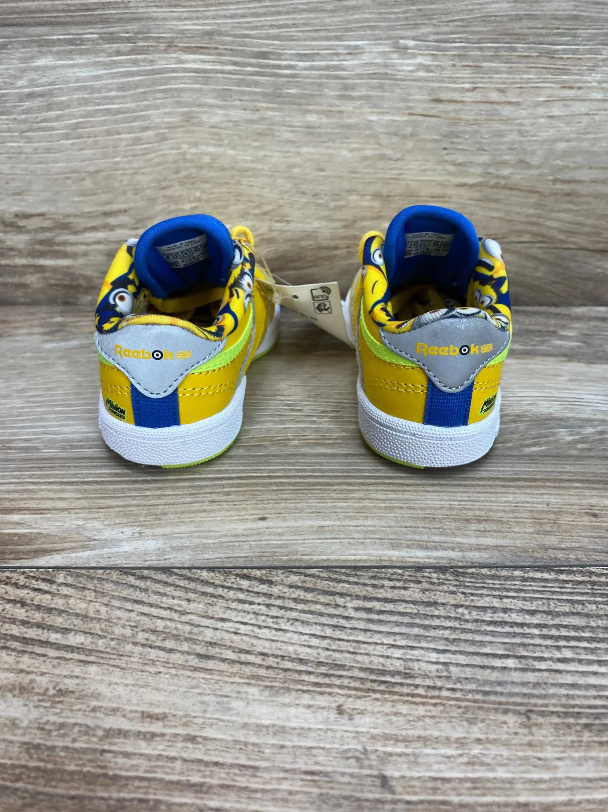 Rear view of toddler Reebok Club C 85 Minions sneakers, yellow and blue with Minions printed collar on wood floor