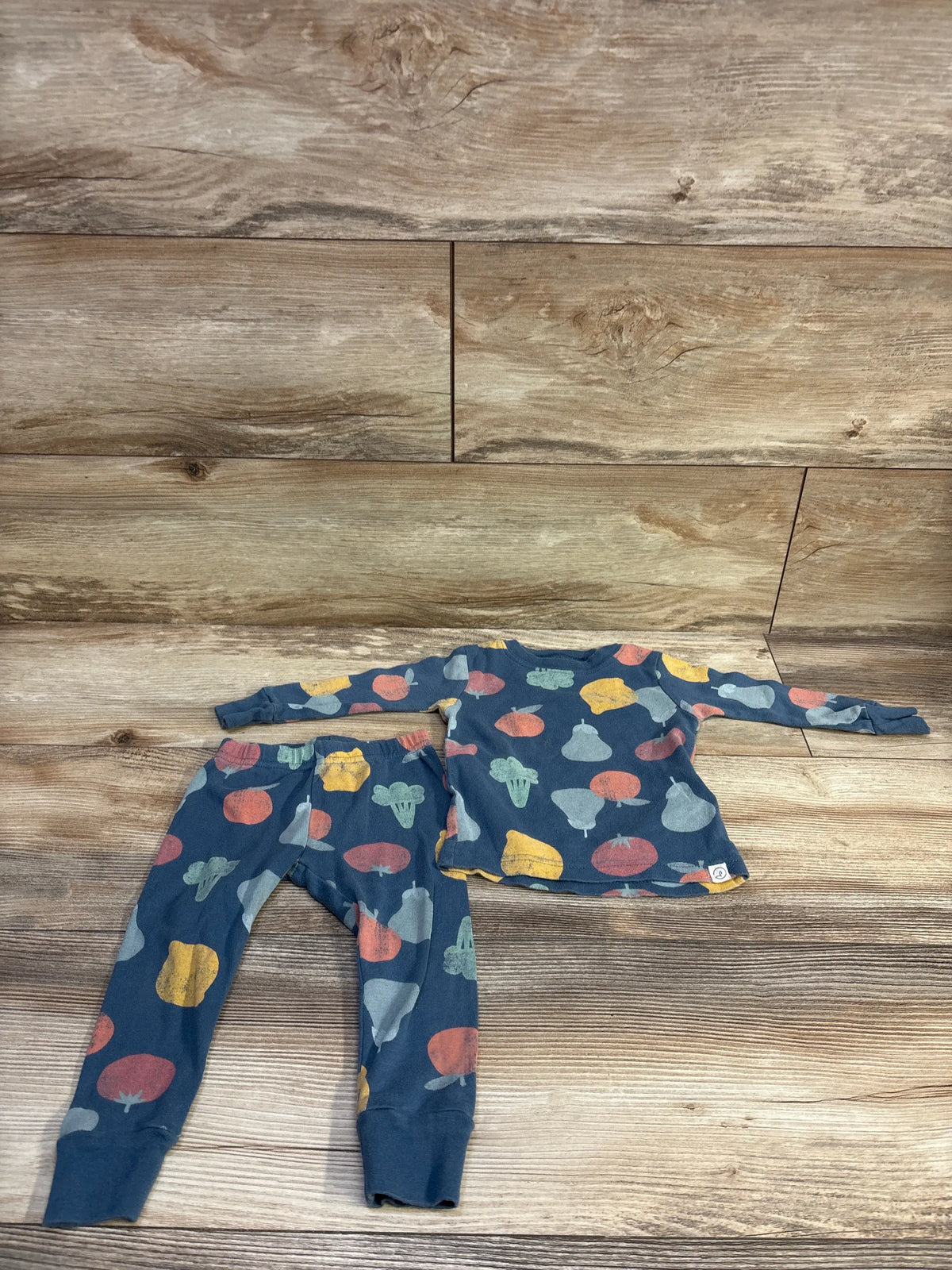 Flat lay of Little Planet Organic blue 2-piece toddler pajama set showing colorful produce print on wood background