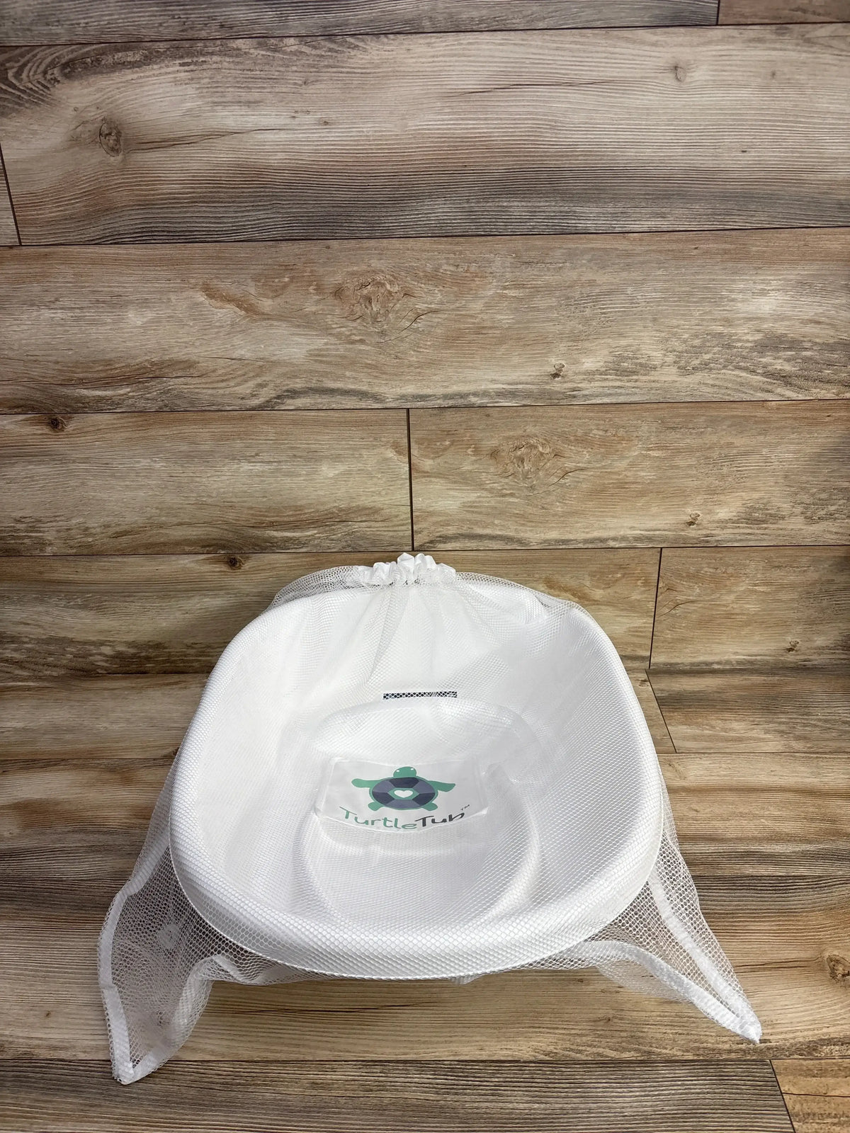 Top-down view of TurtleTub Swaddle Bath Tub showing white tub with mesh swaddle and turtle logo on wood plank floor