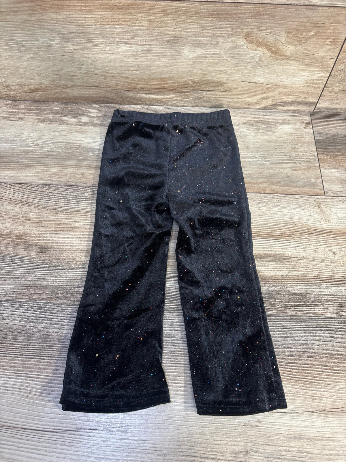 Front view of Tahari black sparkly velvet leggings showcasing multicolor sparkle detailing and elastic waistband on a wood look floor