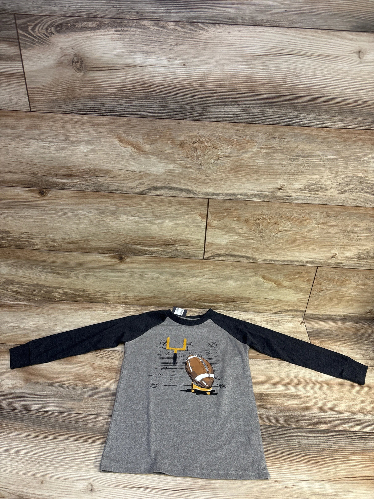 Top-down view of toddler grey long-sleeve raglan shirt showcasing a printed football and goalpost graphic on wood plank background