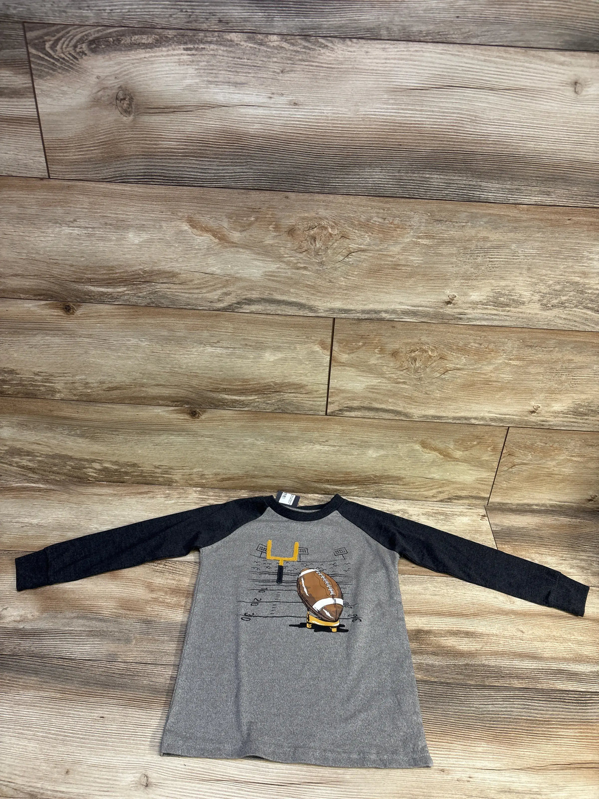 Top-down view of toddler grey long-sleeve raglan shirt showcasing a printed football and goalpost graphic on wood plank background