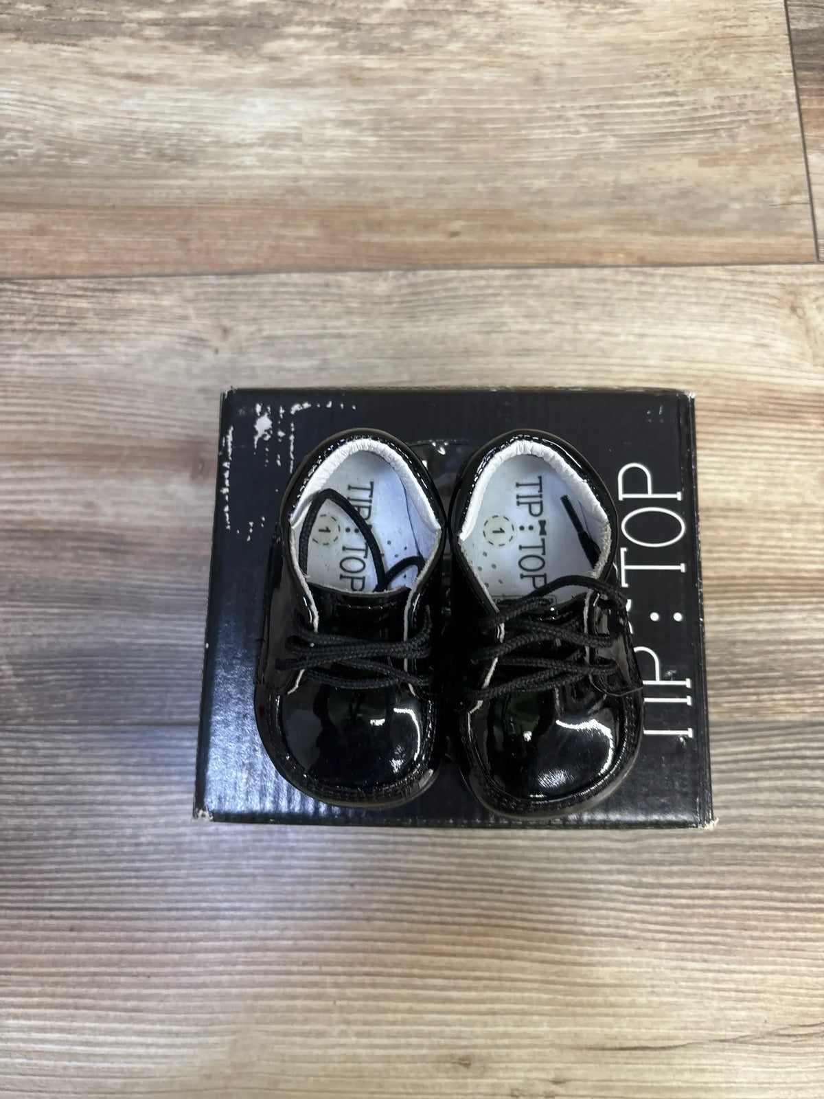 Top down view of Tip Top black patent leather baby sneakers showing glossy finish and laces on original box on wood floor