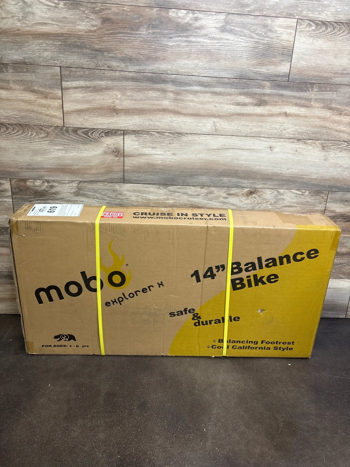 Front view of boxed Mobo Explorer X 14-inch balance bike showing original packaging on a wood-look floor