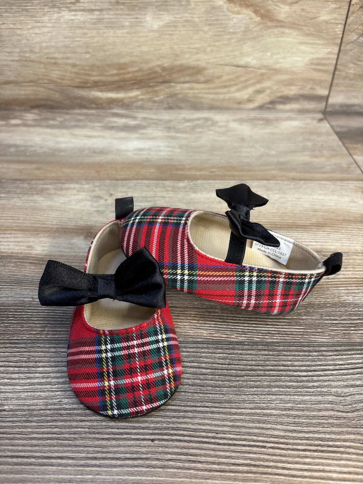 Top view of Little Me soft-sole tartan Mary Jane baby shoes showcasing red plaid fabric and black bows on wood background
