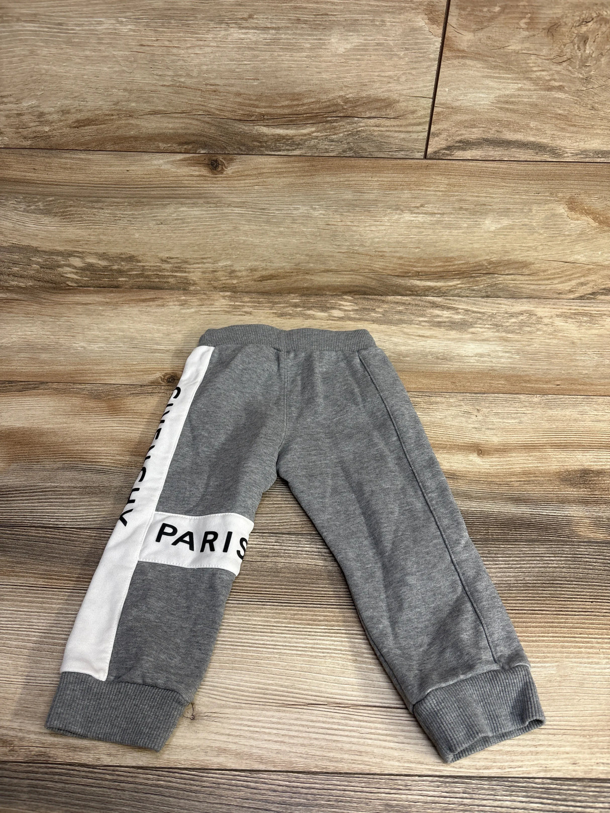 Gray toddler sweatpants with white side panel and black text 'Paris' and 'Cutie City' on wooden floor
