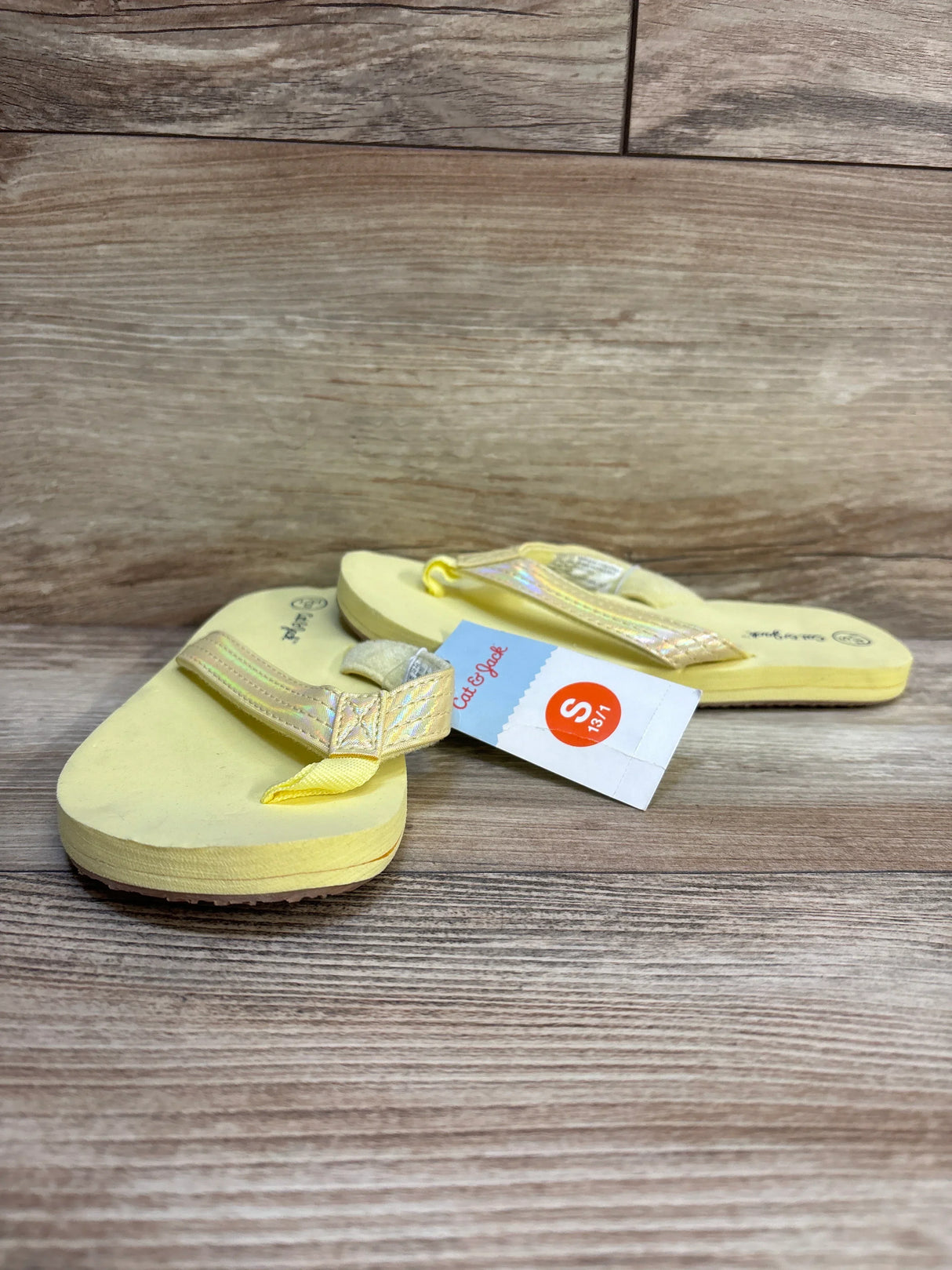 Angled low front view of Cat and Jack Ava toddler flip-flops, yellow with iridescent sparkly straps and visible size tag on wooden floor