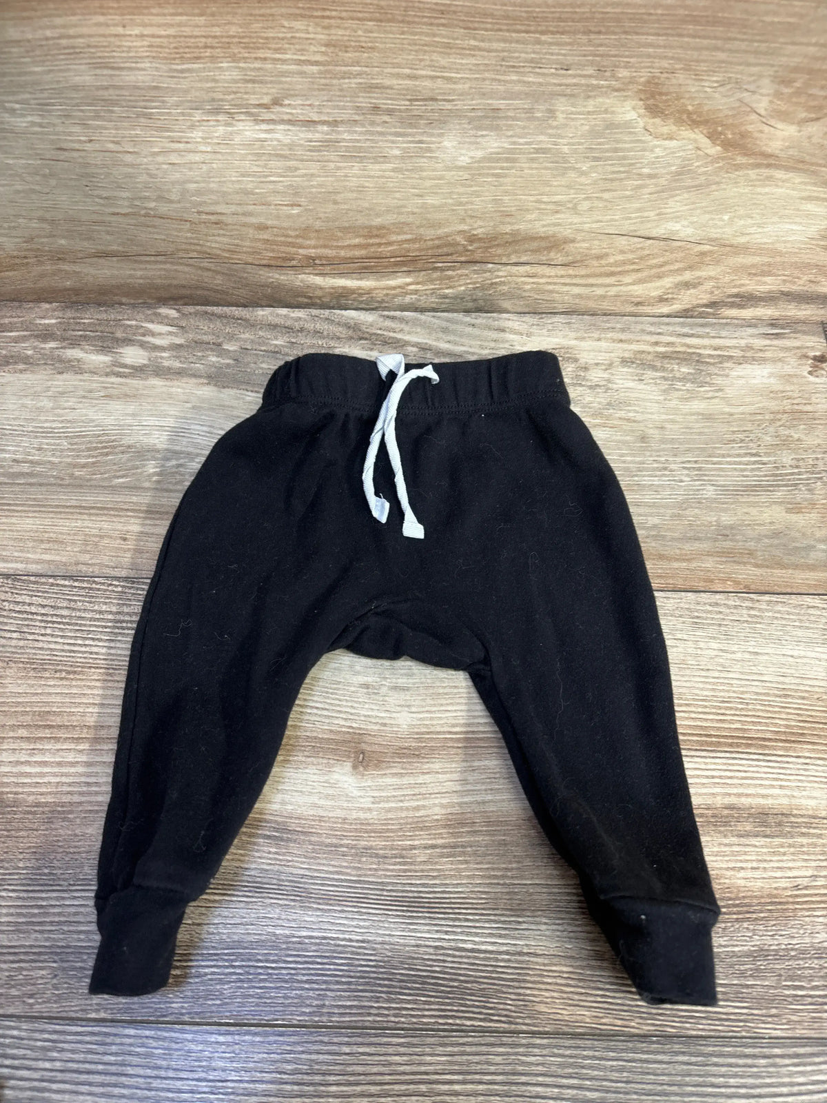 Front view of black drawstring pants for infants showcasing the adjustable drawstring waist and relaxed fit on a wood-grain background