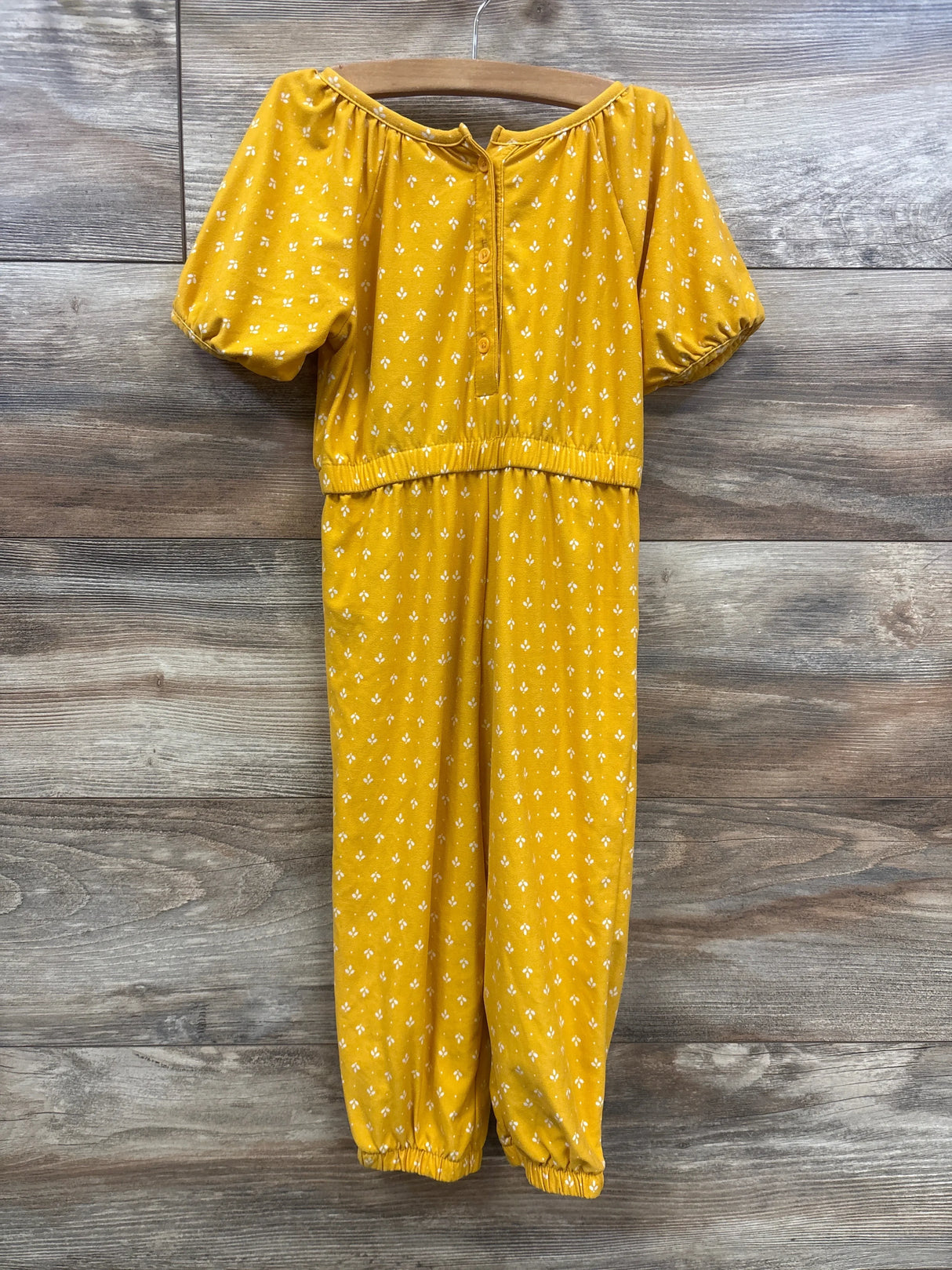 Front of yellow floral girls jumpsuit size 3T with puff sleeves, button front, elastic waist, ankle cuffs on wood background
