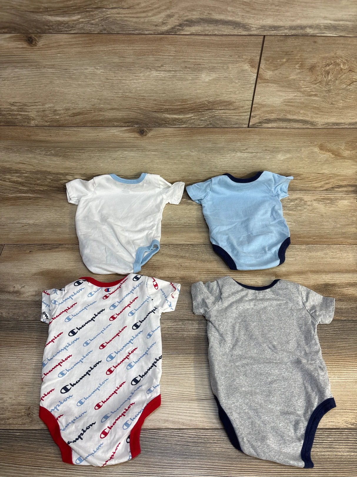 Four baby onesies including Champion print, white, light blue, and gray, laid on wood floor