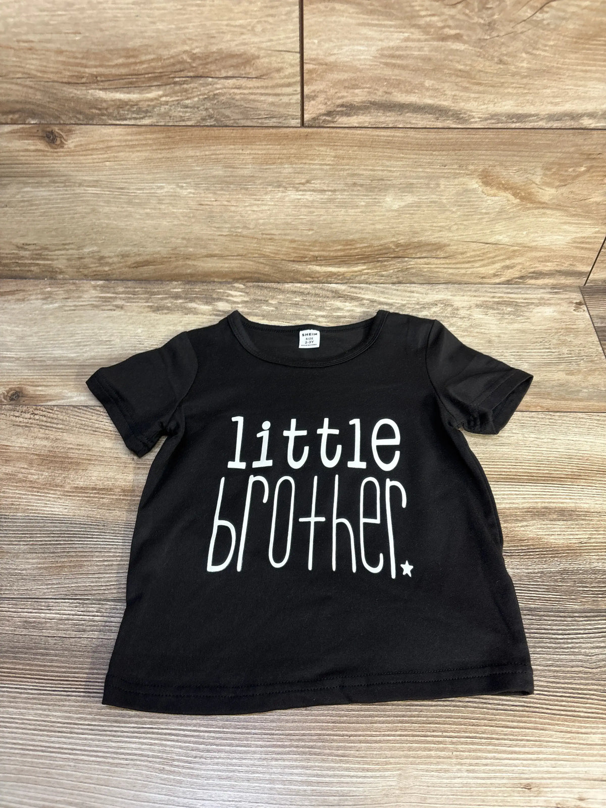 Front view of Shein Little Brother Shirt showcasing white little brother lettering on black fabric with a wood background