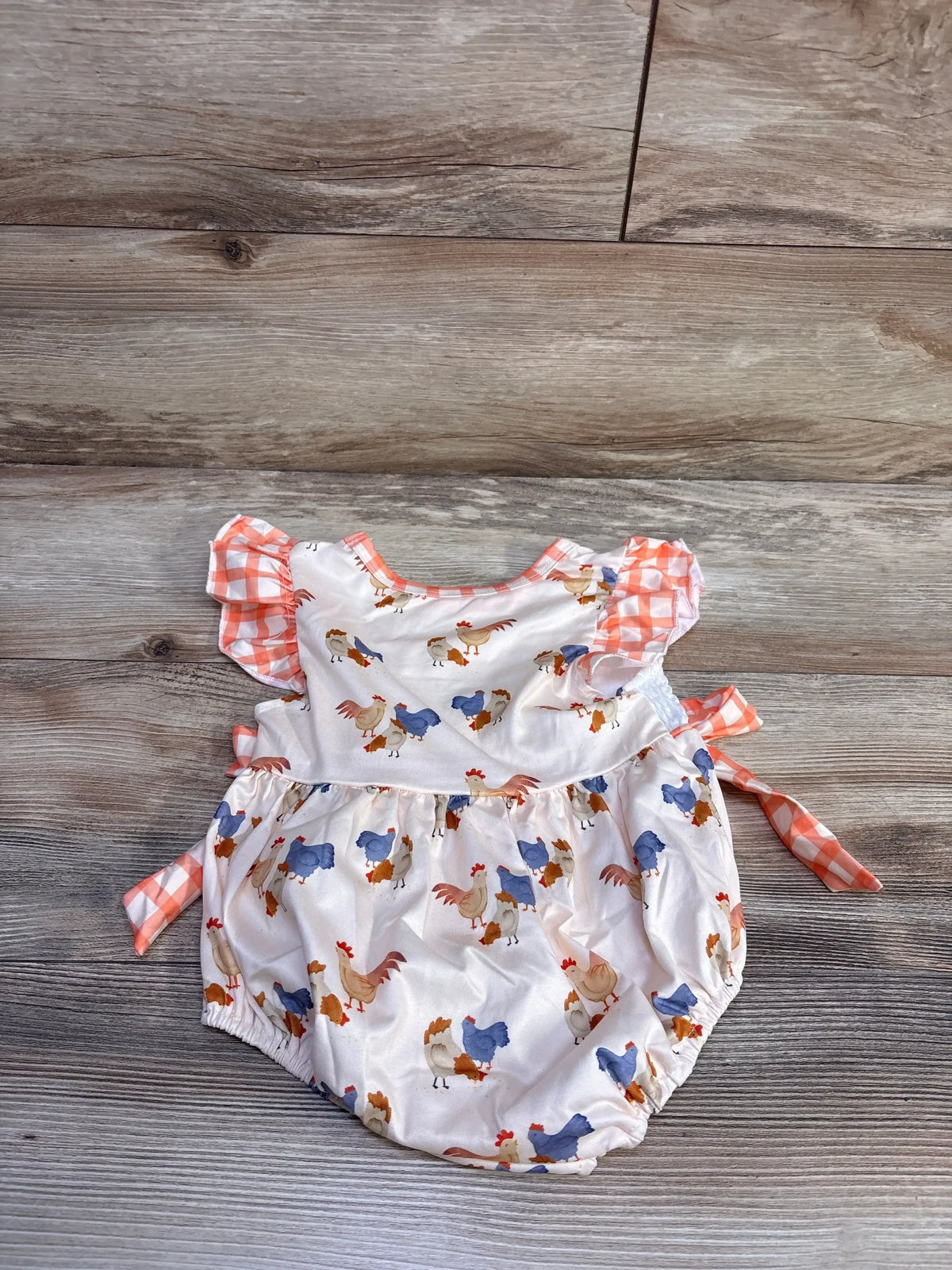 Flat lay of ivory baby bubble romper with rooster print, orange gingham ruffle sleeves and side ties on wood background