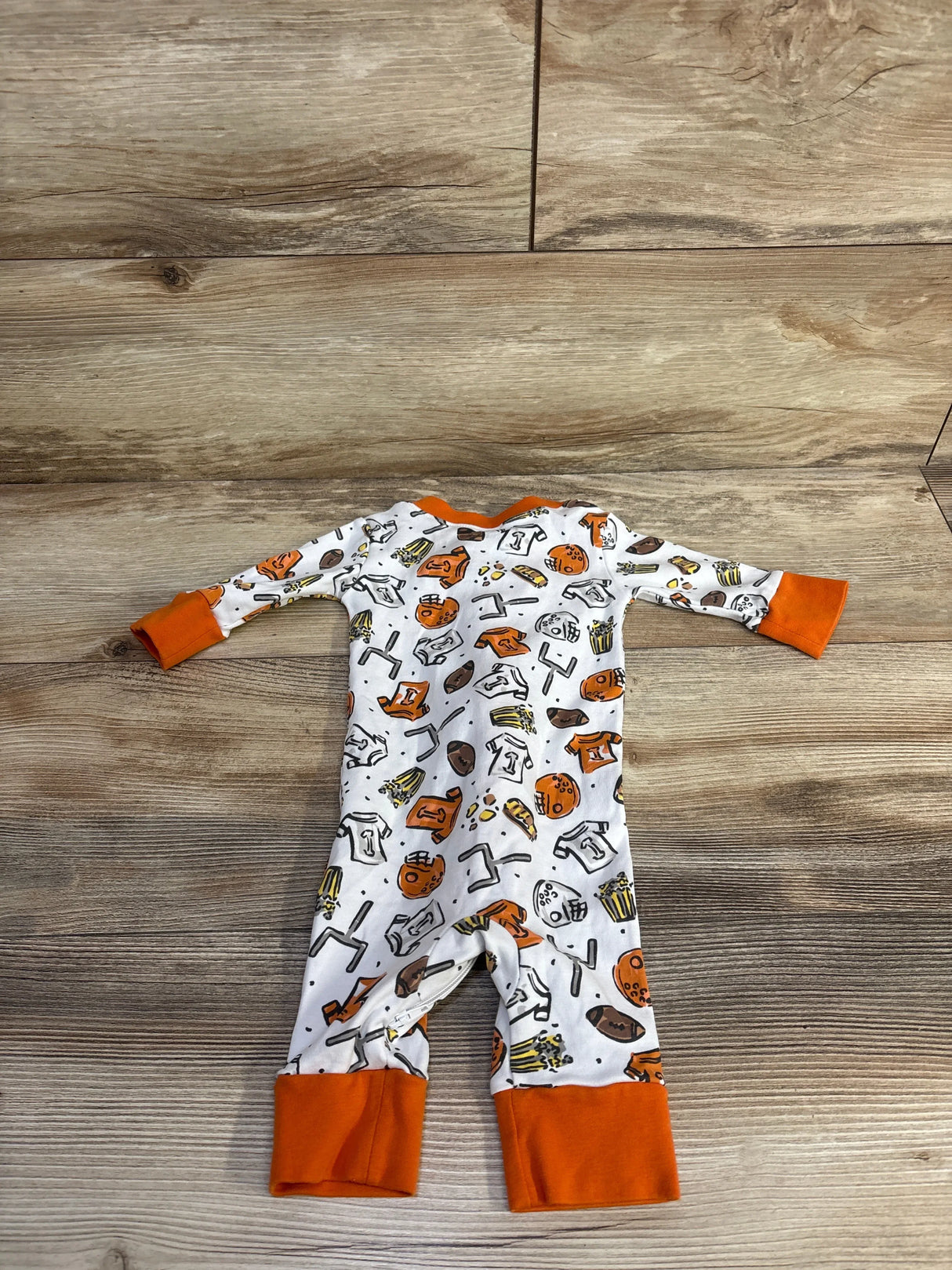 Flat lay of Magnolia Baby footless sleeper size 6 months showcasing orange-trimmed football print and fold-over cuffs on wood background
