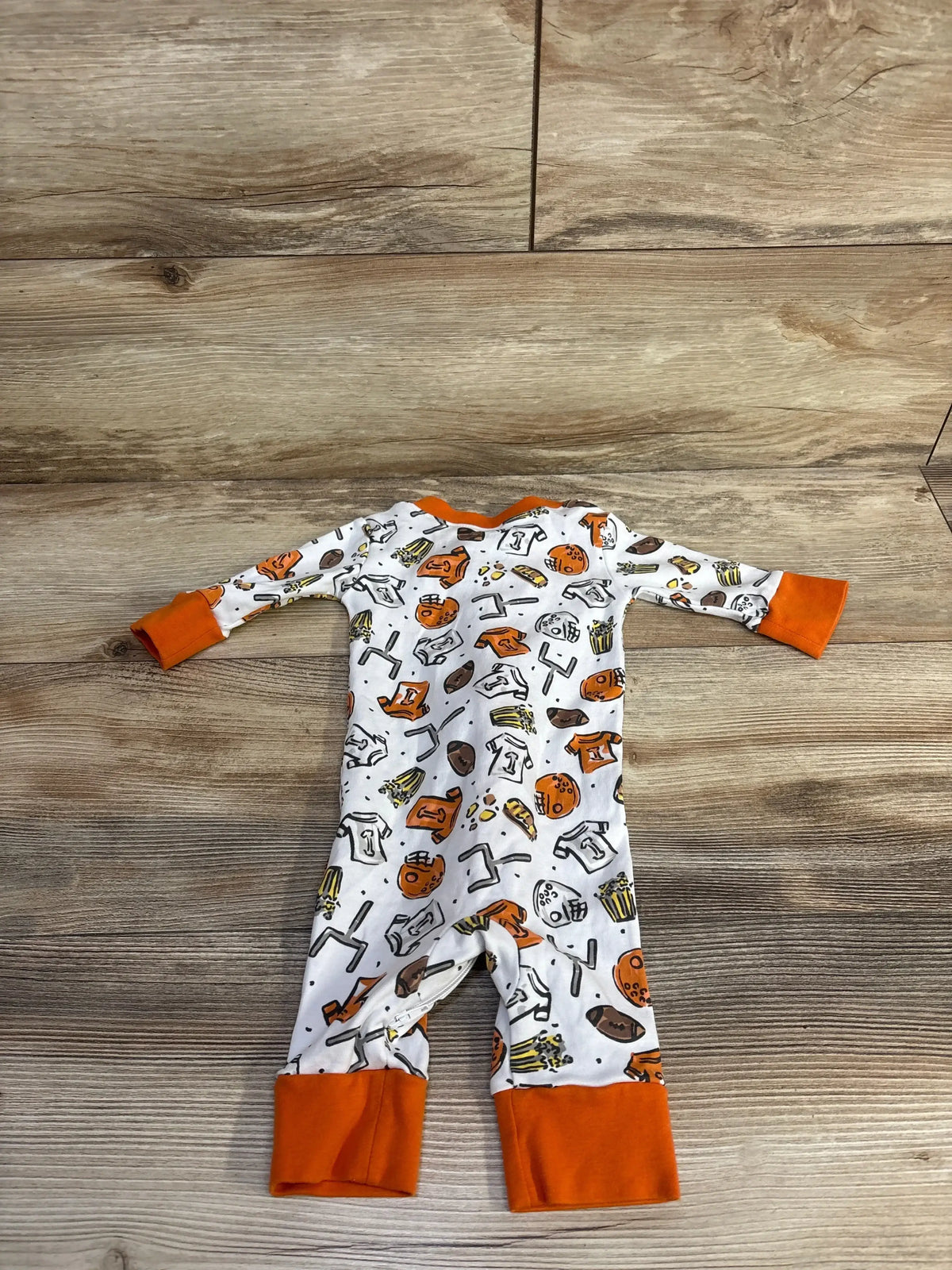 Flat lay of Magnolia Baby footless sleeper size 6 months showcasing orange-trimmed football print and fold-over cuffs on wood background