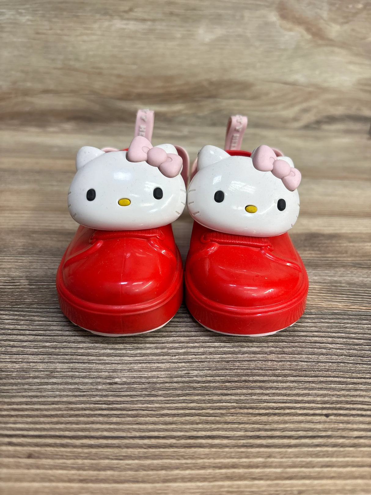 Front view of Mini Melissa BE Hello Kitty red slip-on shoes, size 5C, featuring white Hello Kitty face charms with pink bows on wooden floor