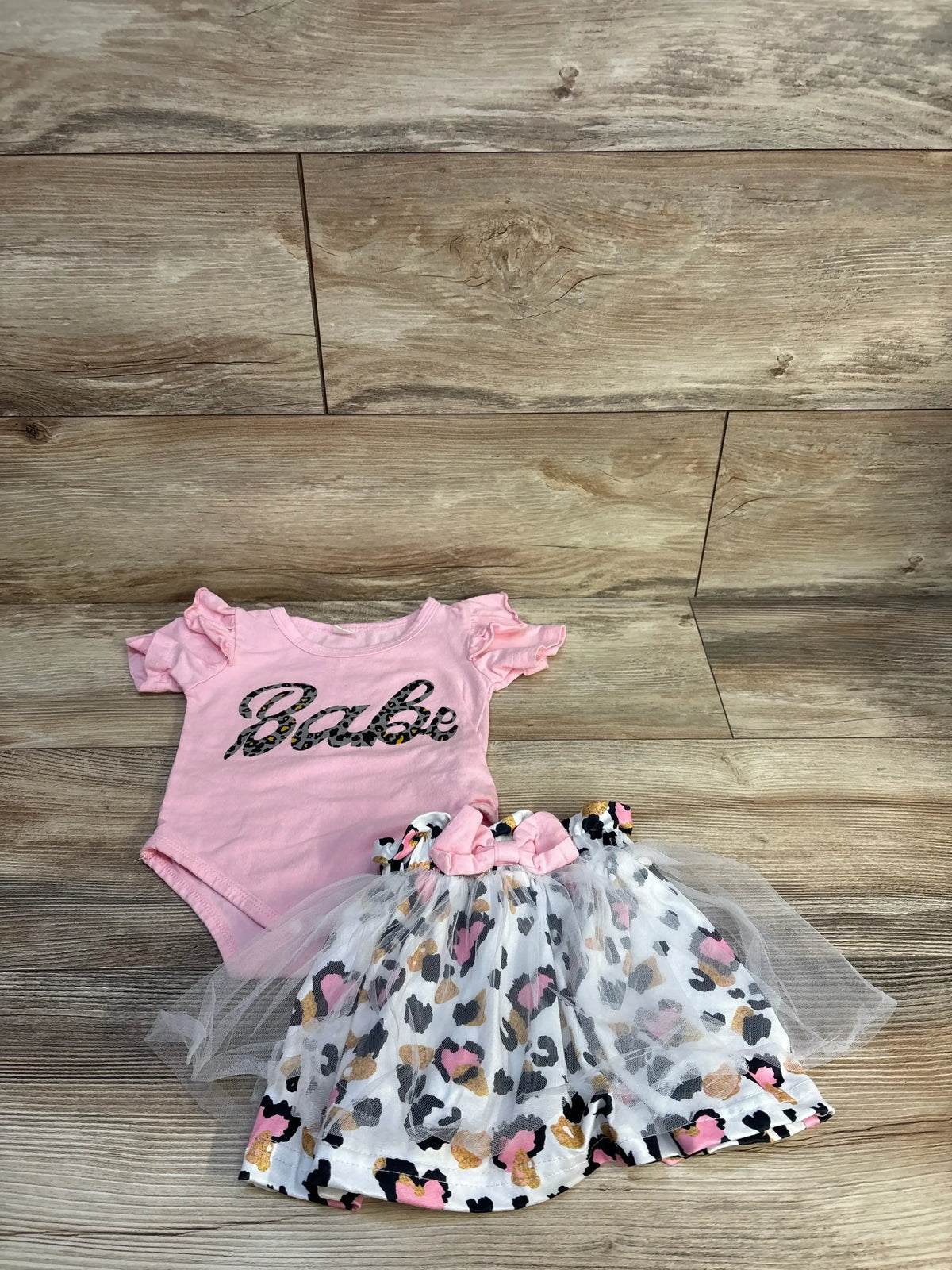 Flat lay of pink Babe bodysuit and leopard print tulle skirt set showcasing ruffled sleeves and bow on wood background