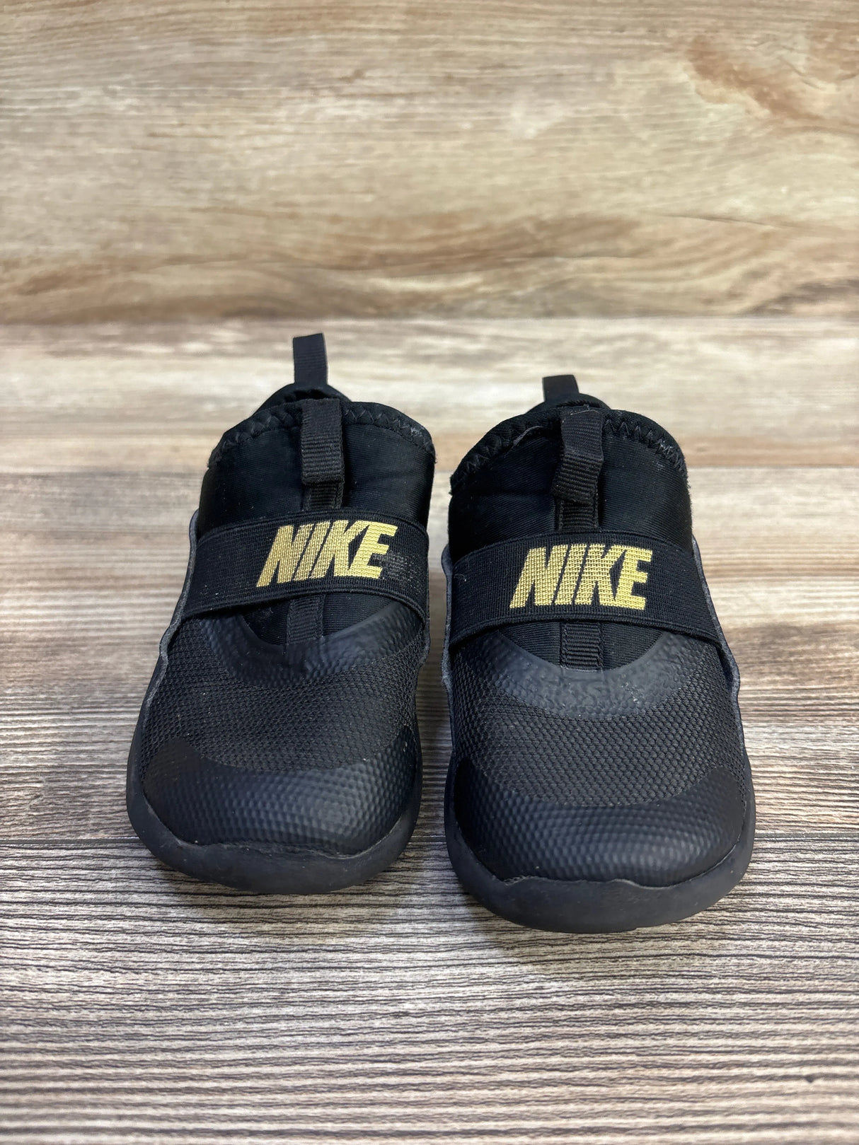 Front view of toddler Nike Flex Runner sneakers, black slip-on with gold NIKE logo on strap, photographed on wood backdrop