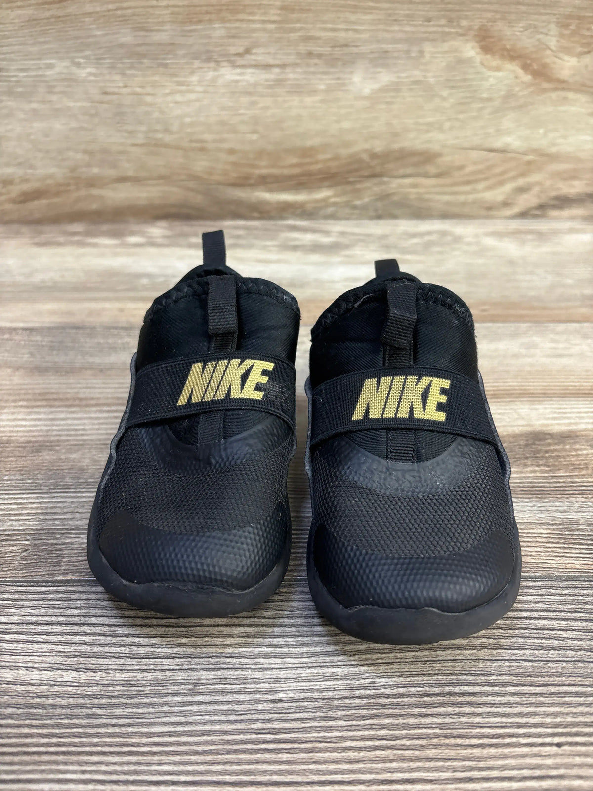 Front view of toddler Nike Flex Runner sneakers, black slip-on with gold NIKE logo on strap, photographed on wood backdrop