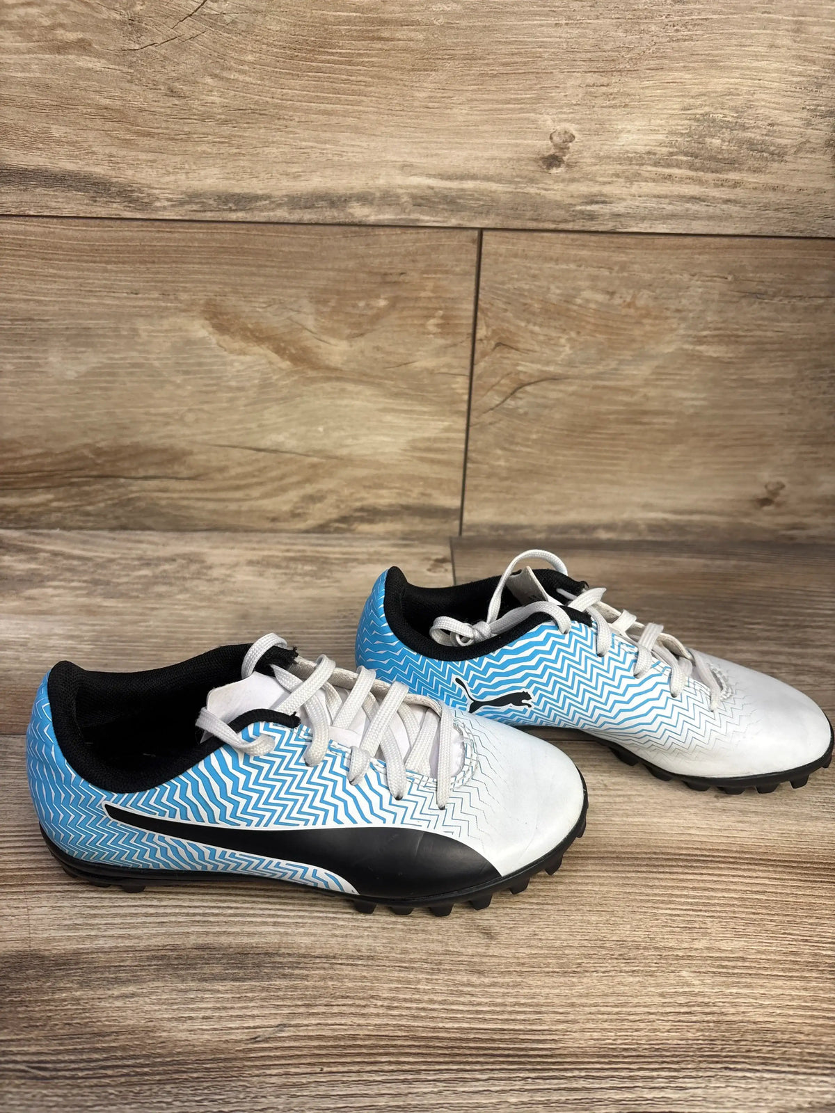 Side view of Puma Rapido II soccer cleats size 12C showing blue zigzag upper, black stripe, and rubber studs on wood floor