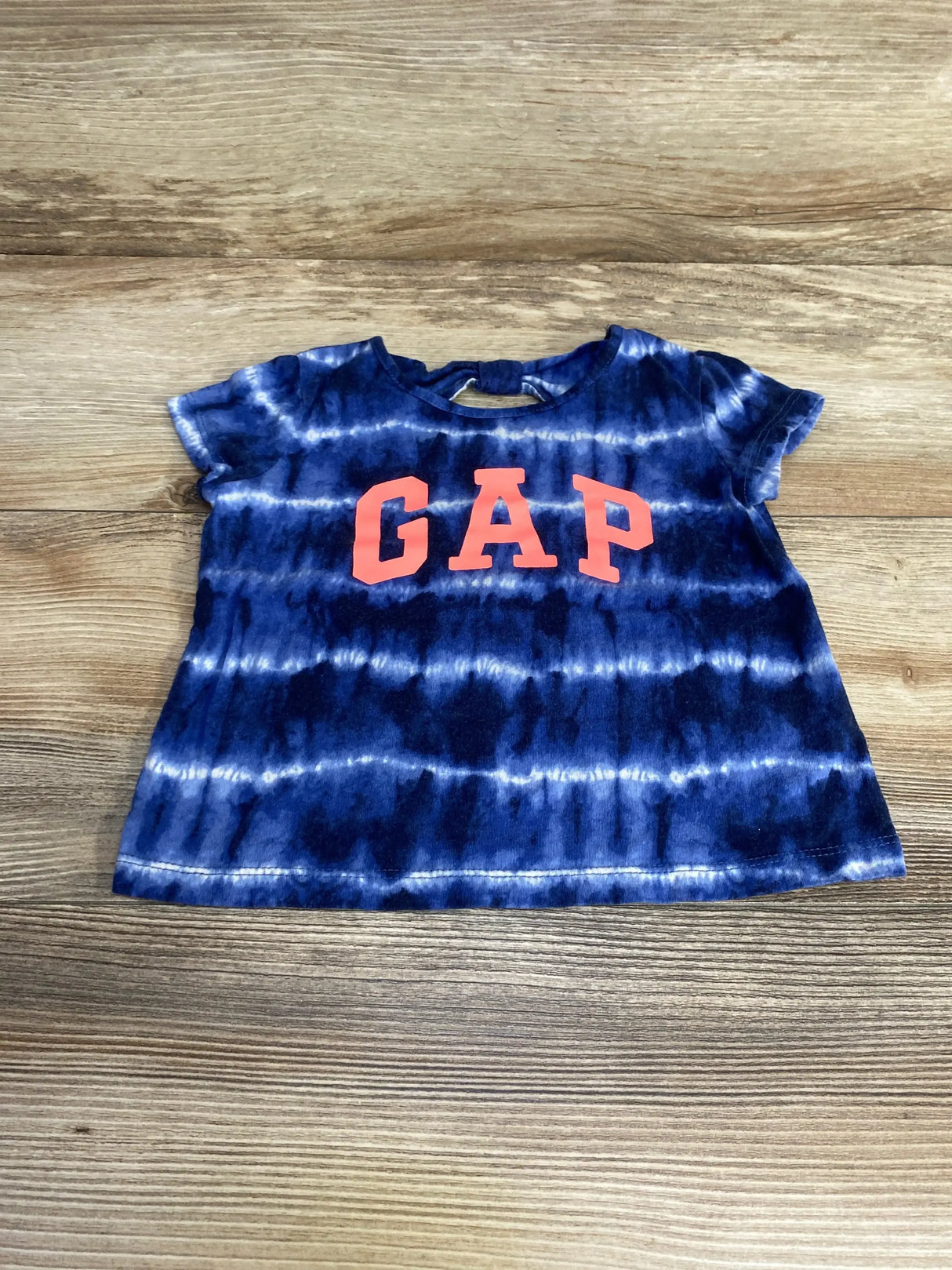 Flat lay of Baby Gap short-sleeve tie-dye shirt showcasing navy and white horizontal stripes and coral GAP letters on wood background