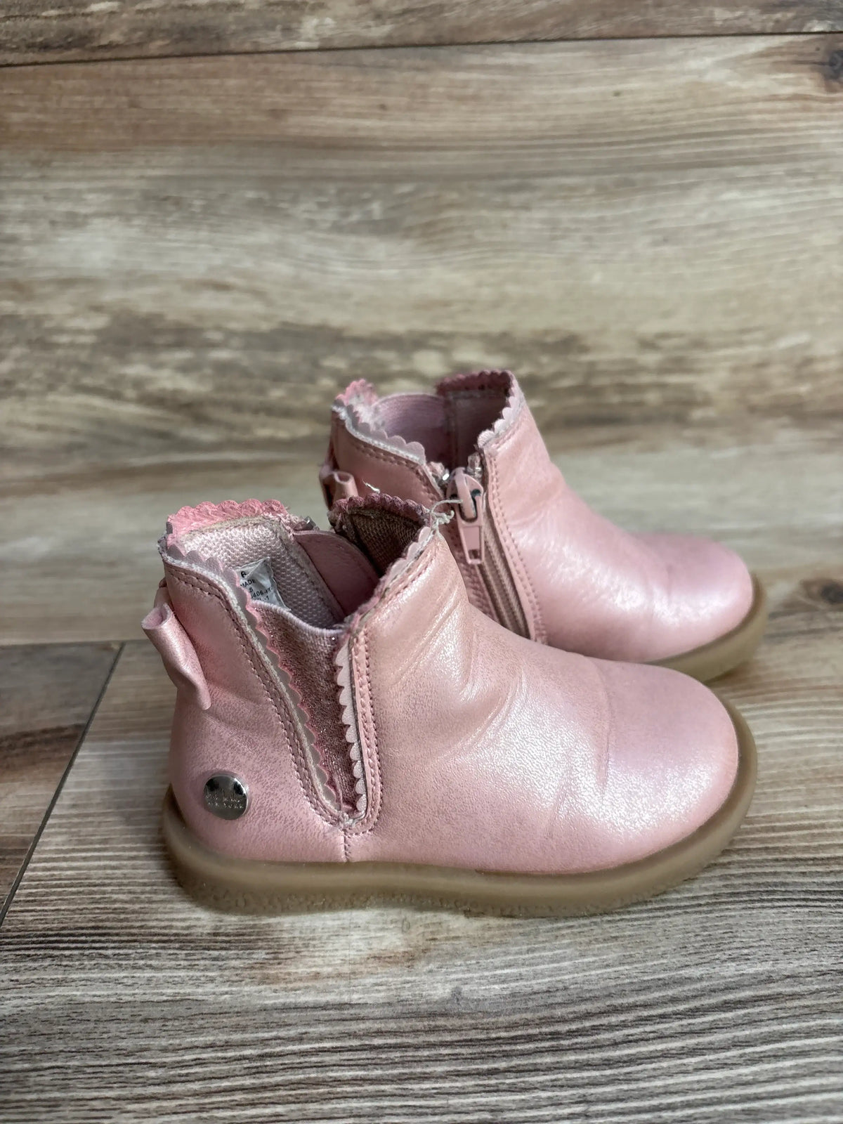 Three-quarter view of Nicole Miller ankle boots size 6c in metallic pink leather with scalloped edges and side zipper on wood plank floor