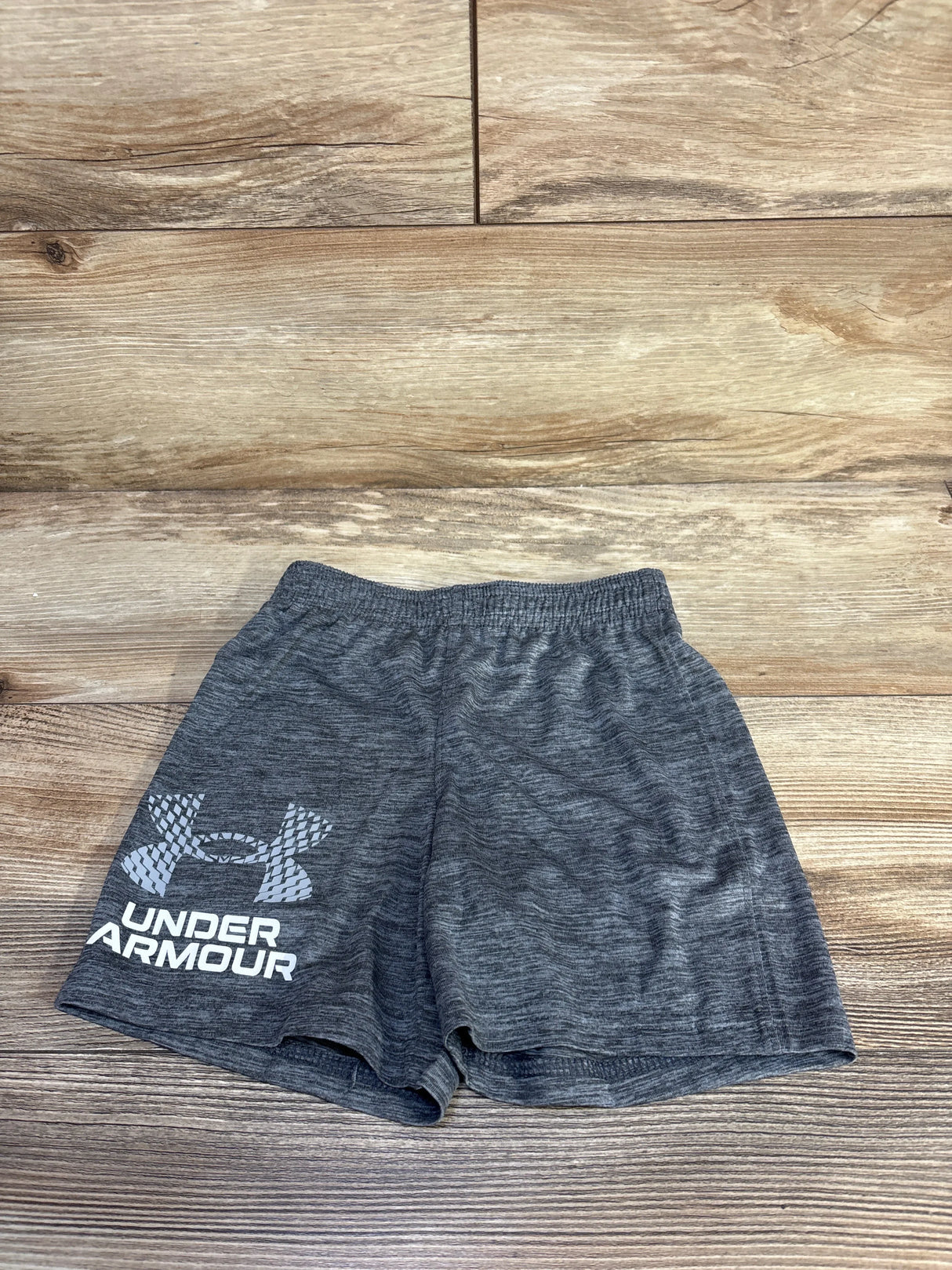 Flat lay of grey Under Armour toddler shorts showing elastic waistband, heathered fabric and large white Under Armour logo on left leg on wood floor