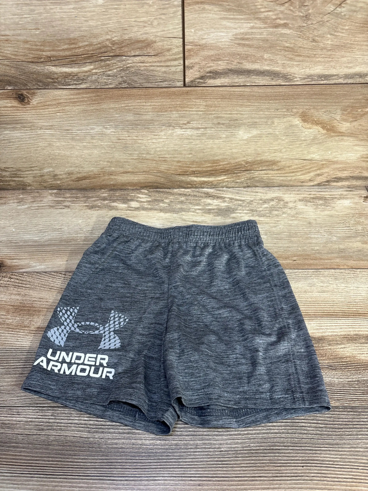 Flat lay of grey Under Armour toddler shorts showing elastic waistband, heathered fabric and large white Under Armour logo on left leg on wood floor