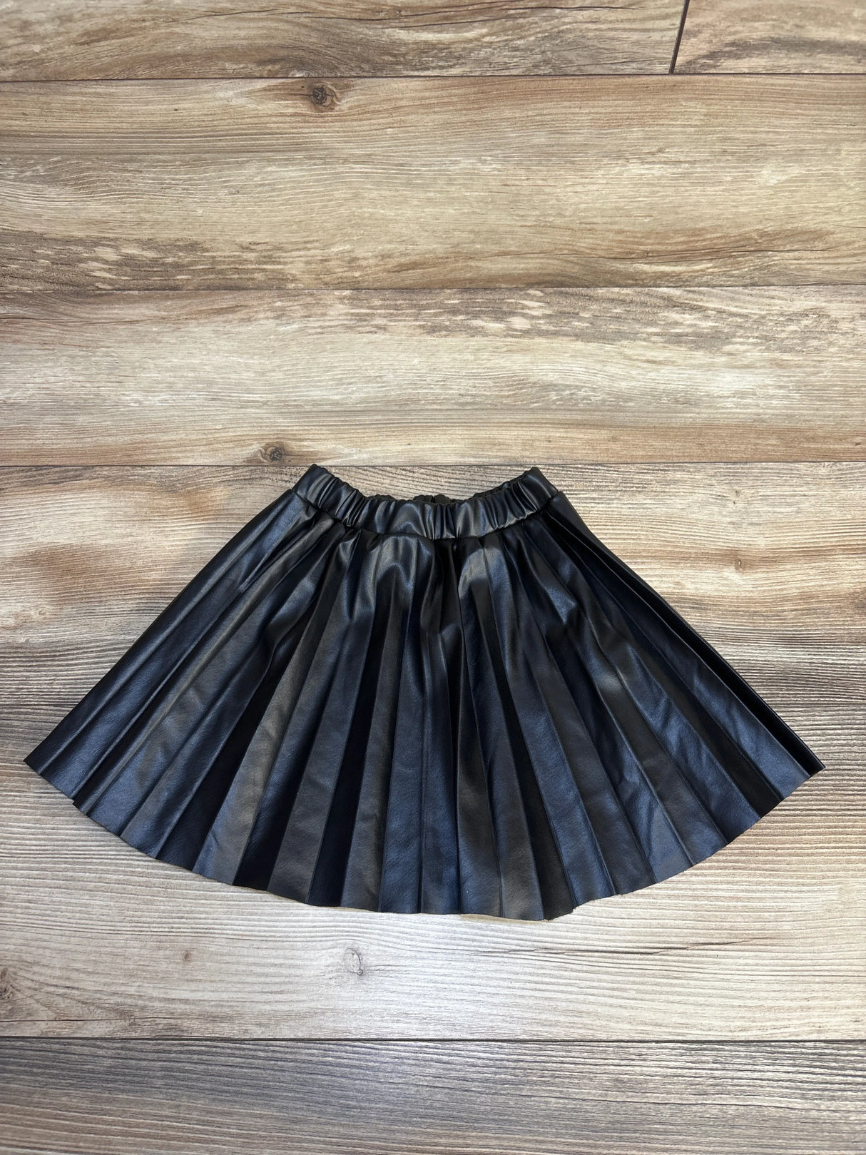 Black pleated faux leather skirt for kids, laid flat on wood floor