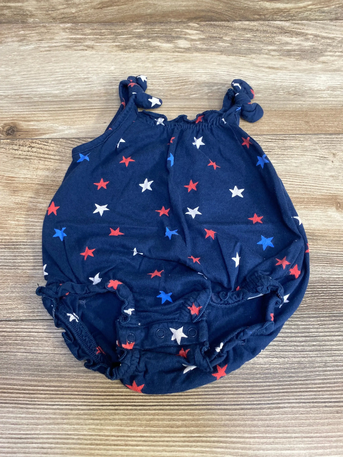 Flat lay of navy Old Navy romper with red white and blue stars, knotted shoulder straps and crotch snaps on wood surface