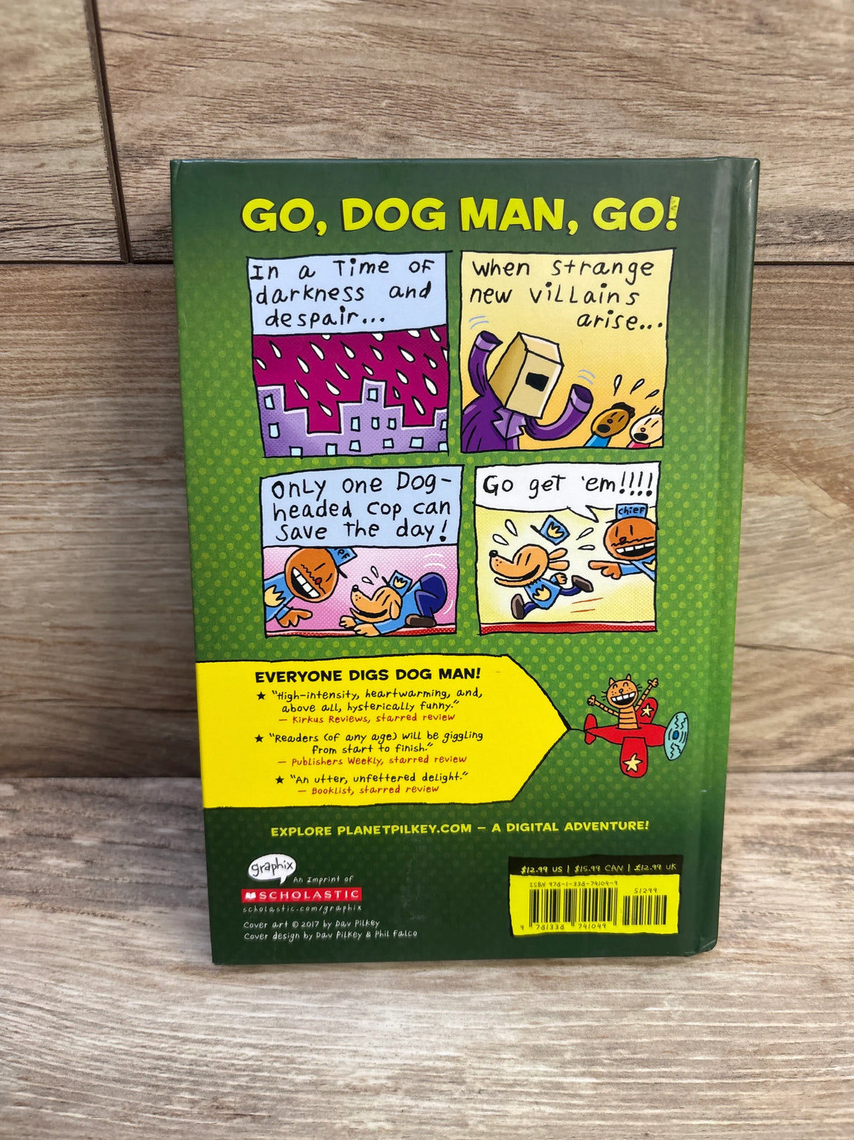 Back view of Dog Man Unleashed #2 hardcover book showing GO, DOG MAN, GO! headline and comic-strip back cover on wooden surface