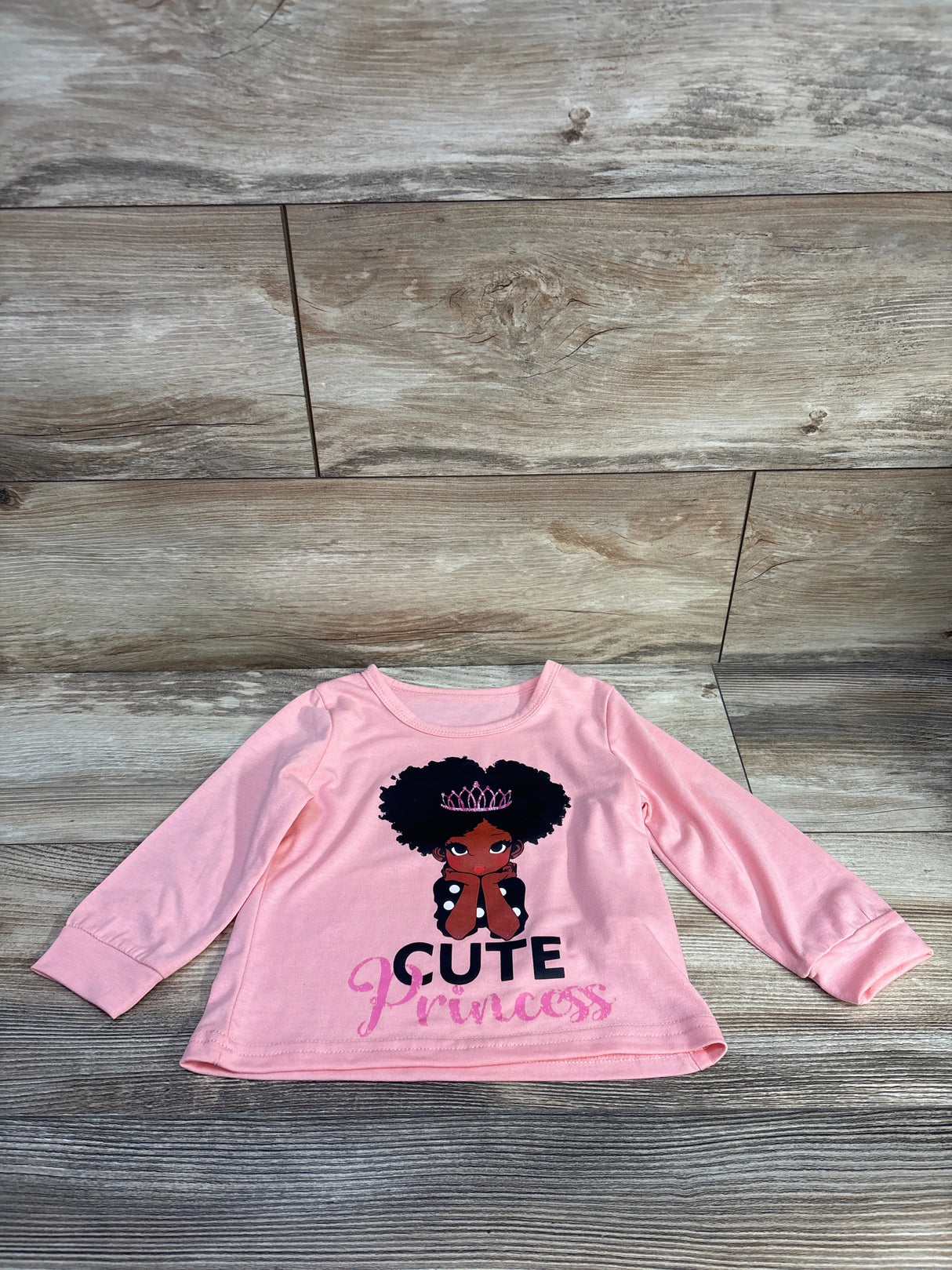Front flat lay of pink girls long sleeve shirt showing Afro girl graphic with tiara and Cute Princess text on wood background