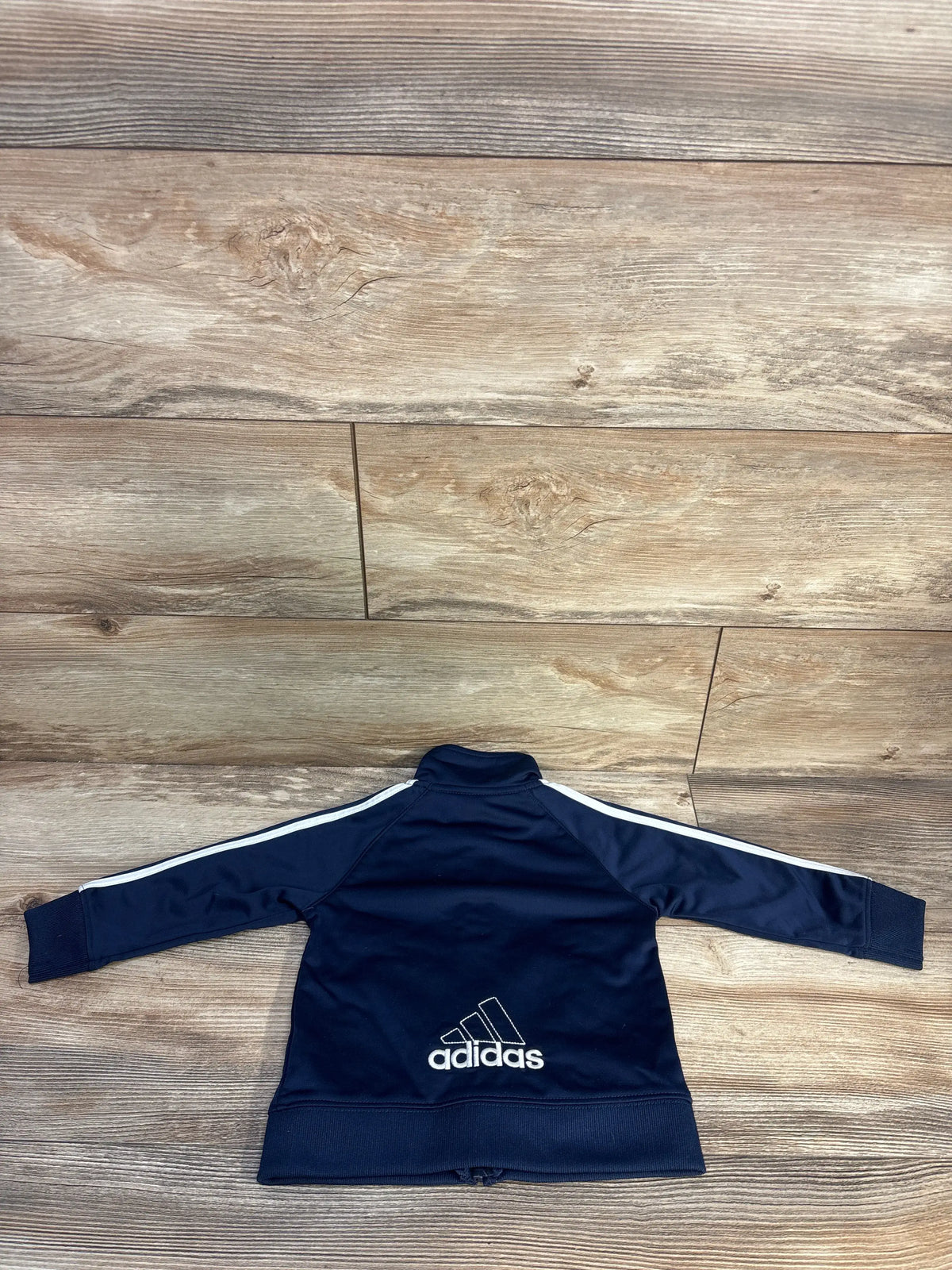 Back view of navy blue Adidas toddler track jacket showing embroidered logo and white sleeve stripes on light wood floor