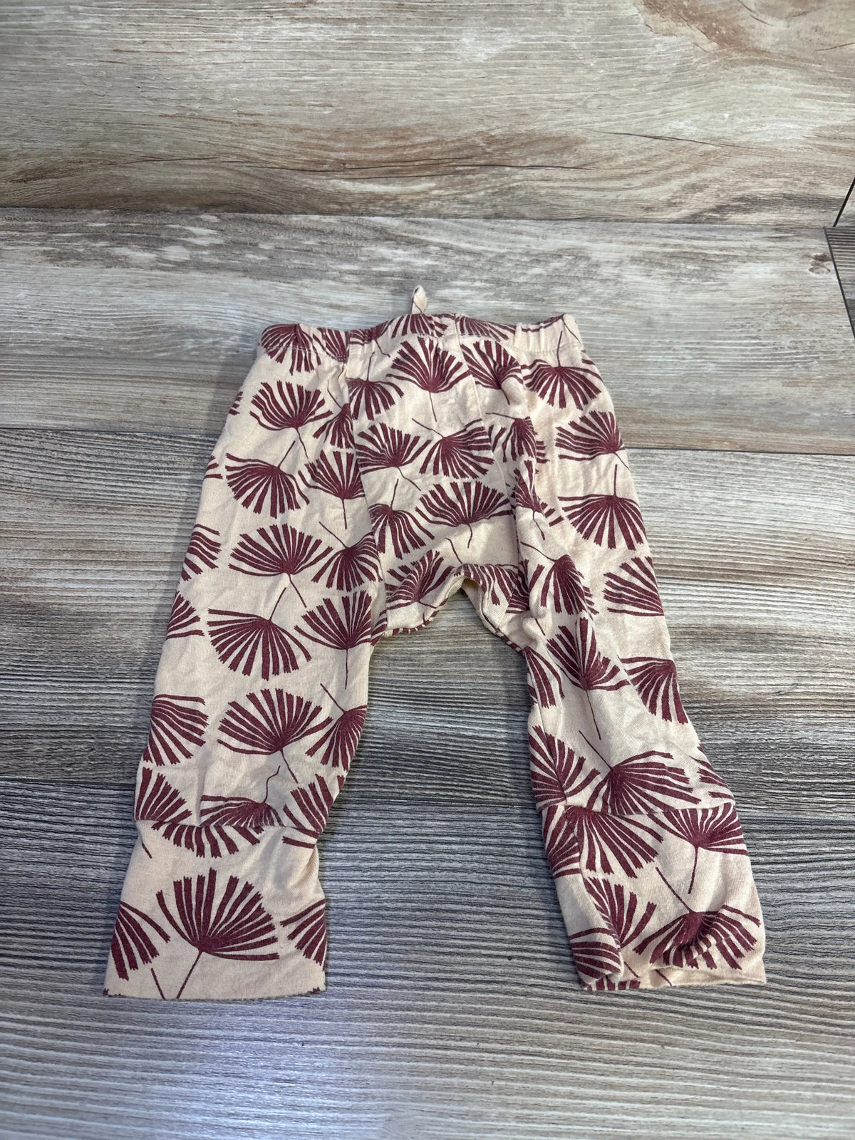 Flat lay of tan baby bamboo pants size 6 to 12 months showcasing maroon palm-leaf print and elastic waistband on wood background