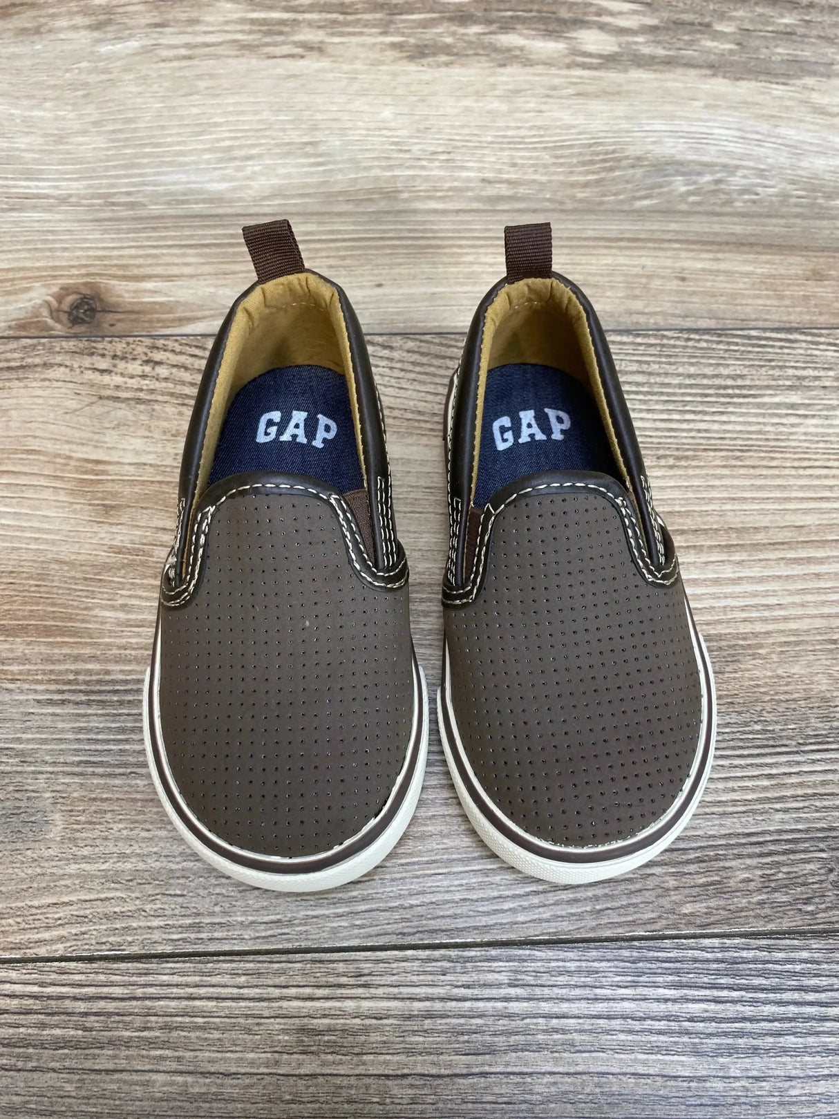 Top view of brown Gap toddler slip-on sneakers with perforated uppers, white soles, contrast stitching, and navy insoles on wood floor