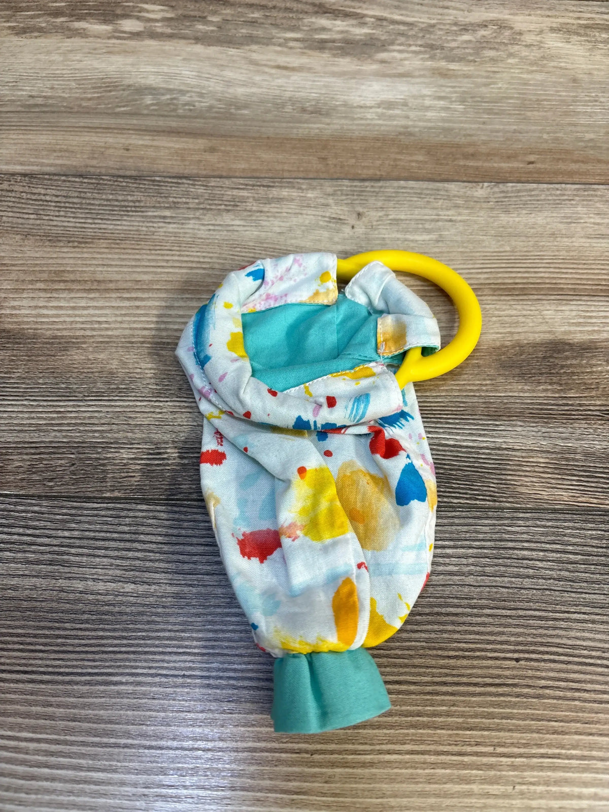 Top view of Lovevery organic cotton sensory pouch with paint-splatter fabric and yellow teething ring on wood surface