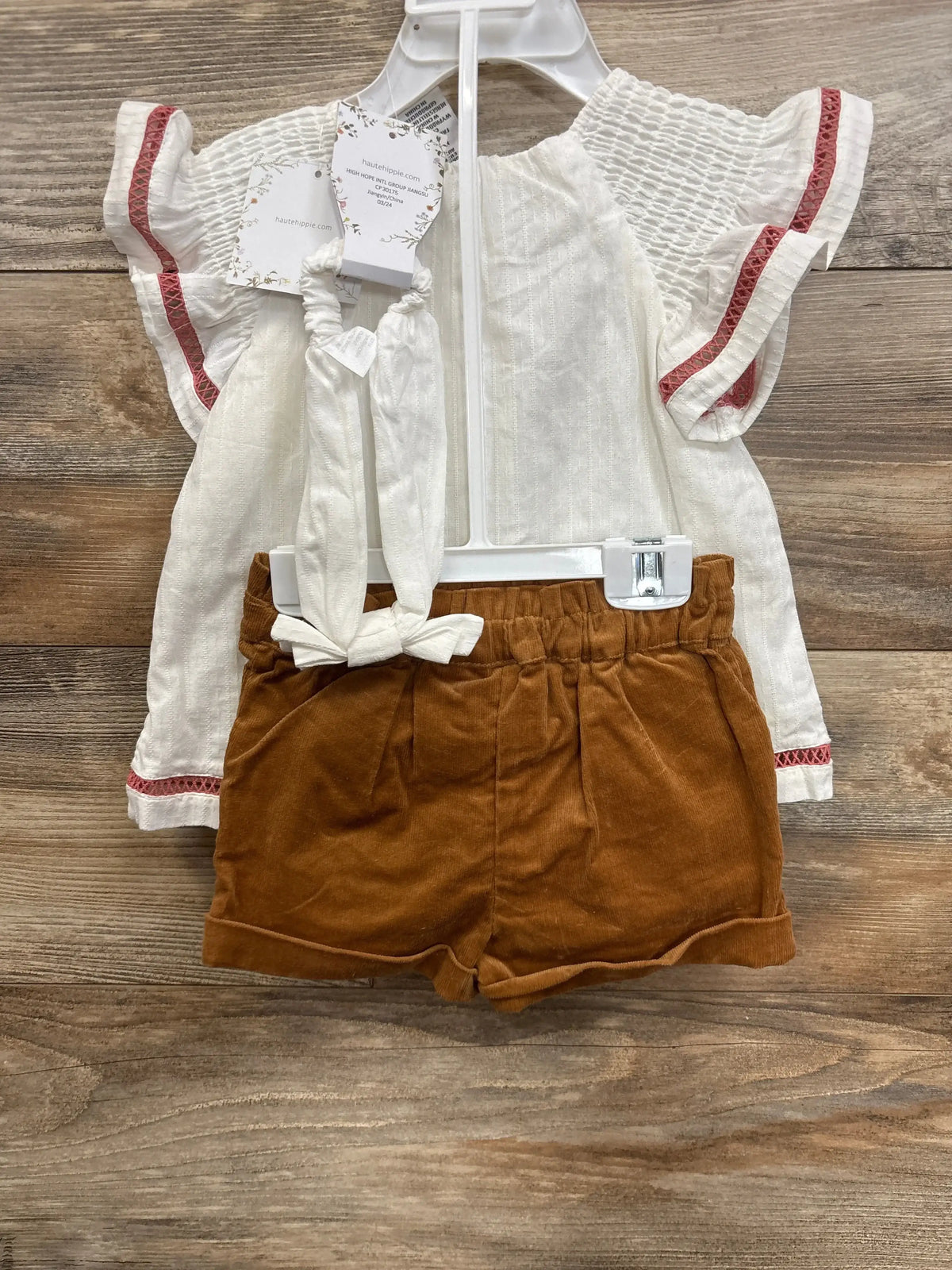 Flat lay of NEW Haute Hippie 3pc layette set showcasing a white embroidered top, corduroy shorts, and headband on wood