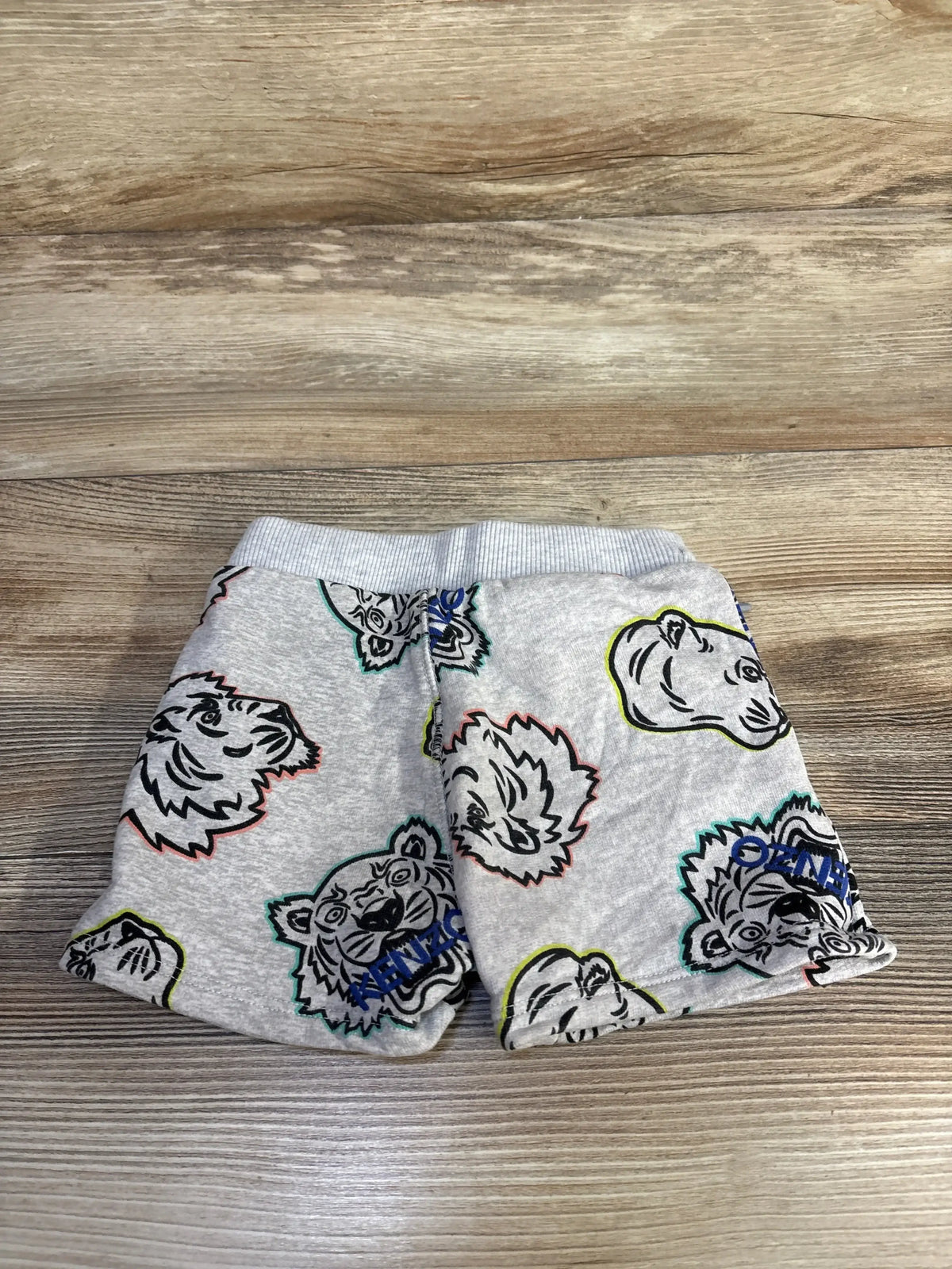 Flat lay of grey Kenzo Kids cotton shorts showcasing multicolored tiger-head sticker print and ribbed elastic waistband on wood background