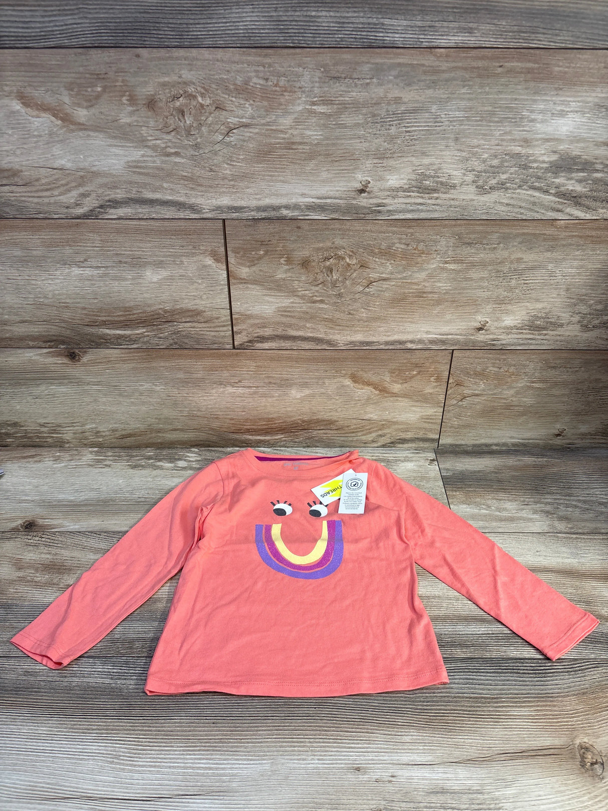 Flat lay of Epic Threads long-sleeve toddler shirt (4T) showcasing coral color and smiling rainbow graphic on wood background